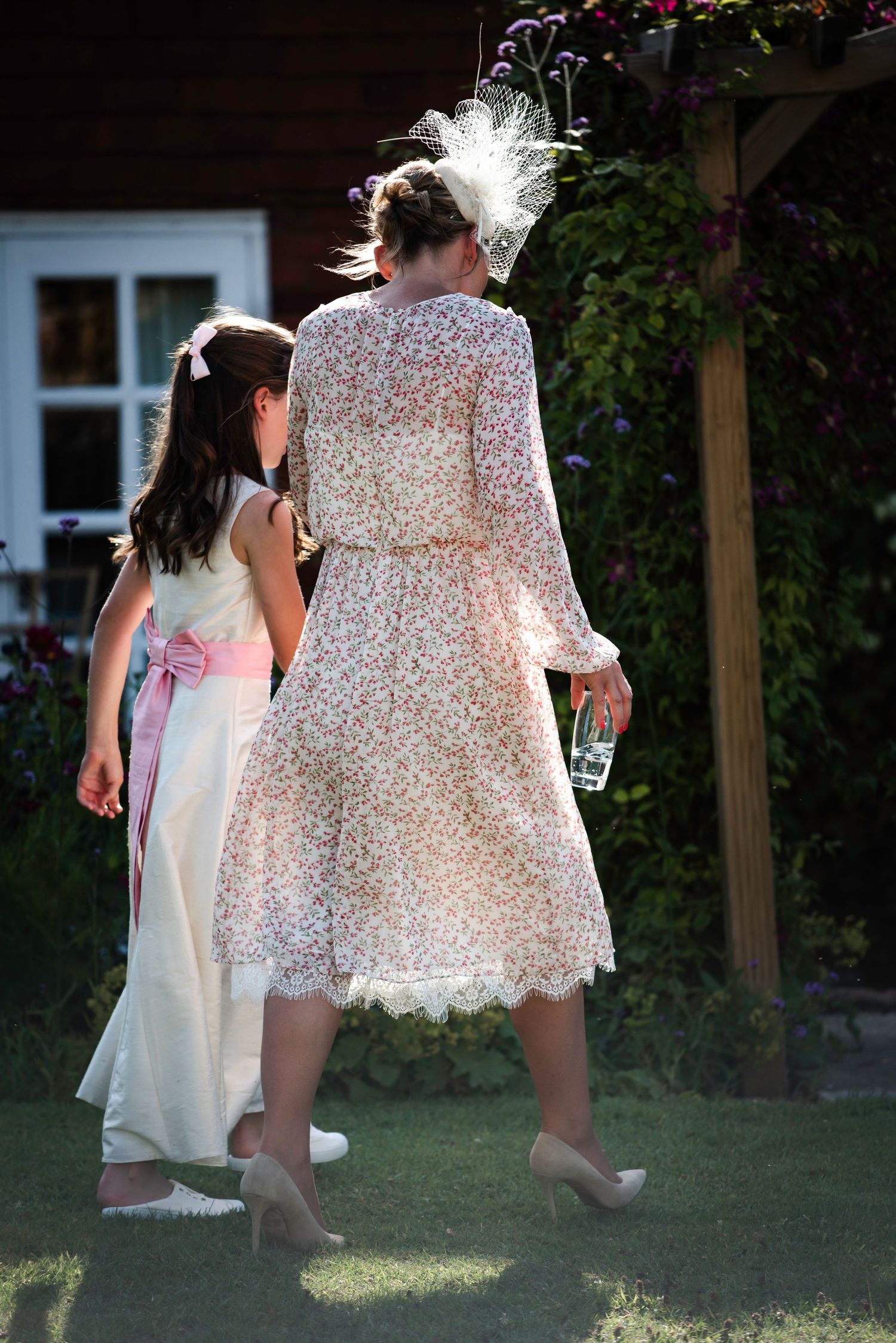 Young flower girl and female wedding guest in floral dress and fascinator walk through garden pergola.