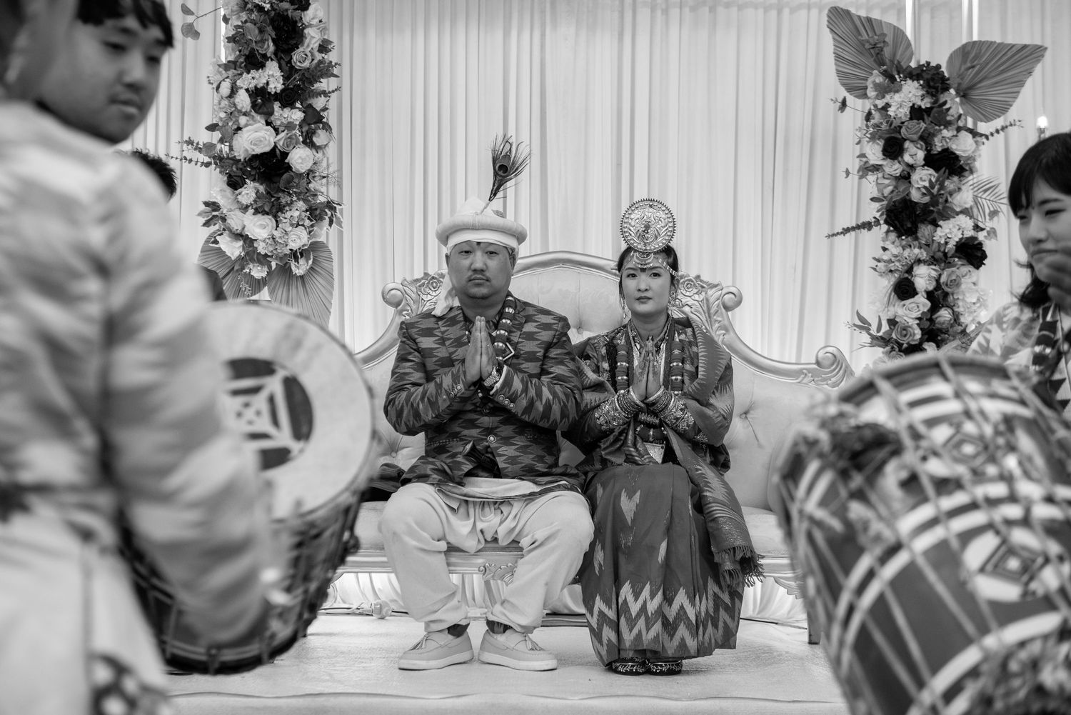 Bride and groom in traditional cultural wedding attire sit on stage with hands in prayer pose.