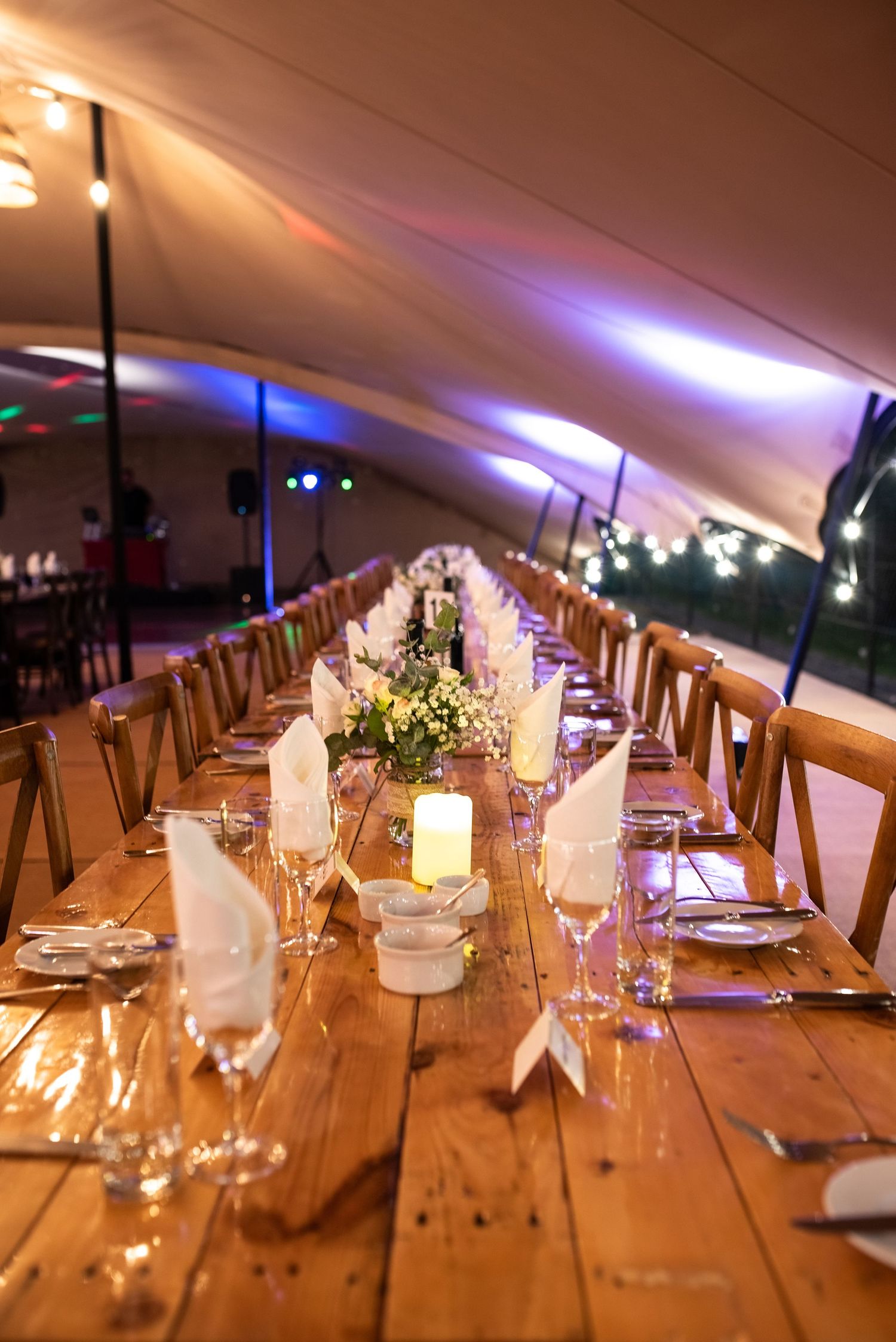 Long rustic wooden banquet table with candles and white napkins under a lit marquee.