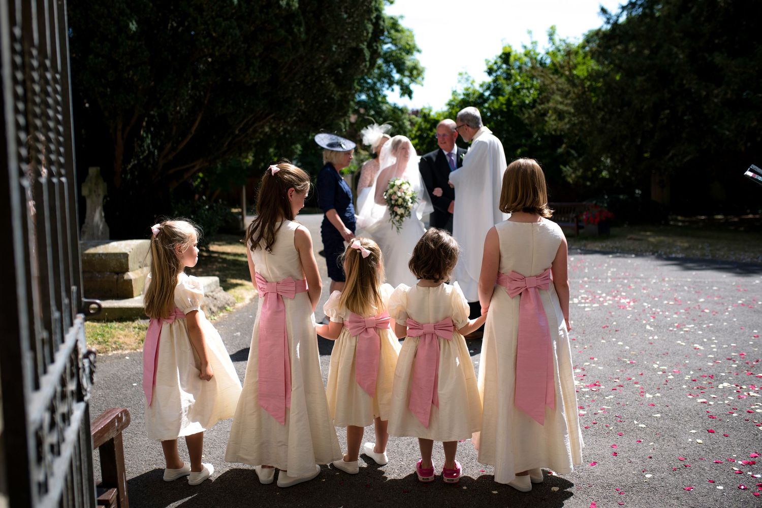 Five flower girls in matching cream and pink dresses stand together watching a wedding ceremony outdoors. St Michaels Church, Bray