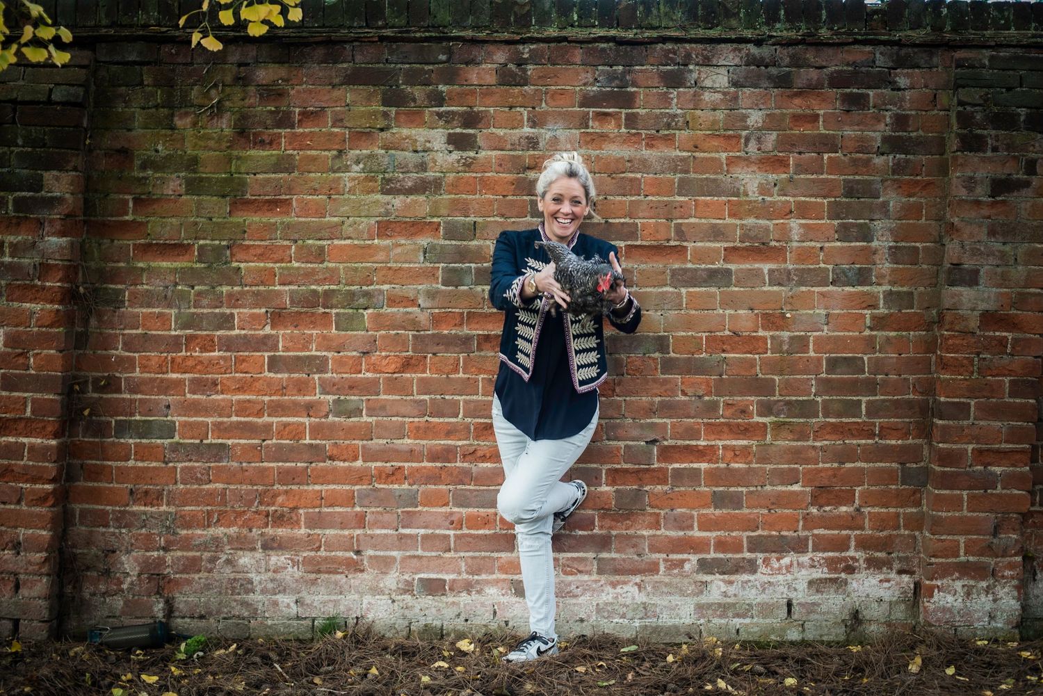 Smiling blonde woman leaning against rustic brick wall holding a phone outdoors.