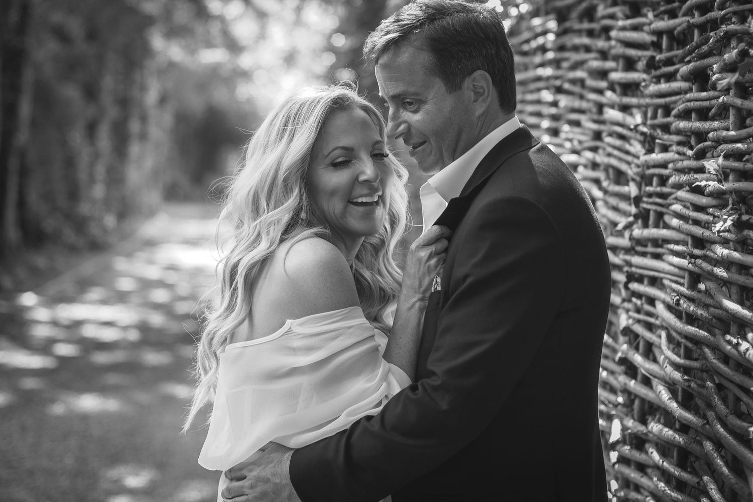 A romantic black and white portrait of a couple embracing beside a wicker fence in dappled light.