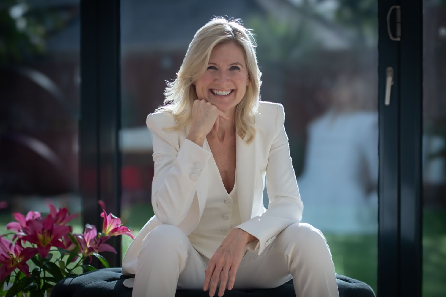 Elegant blonde woman in white suit smiling while seated near pink flowers outdoors.  business portrait