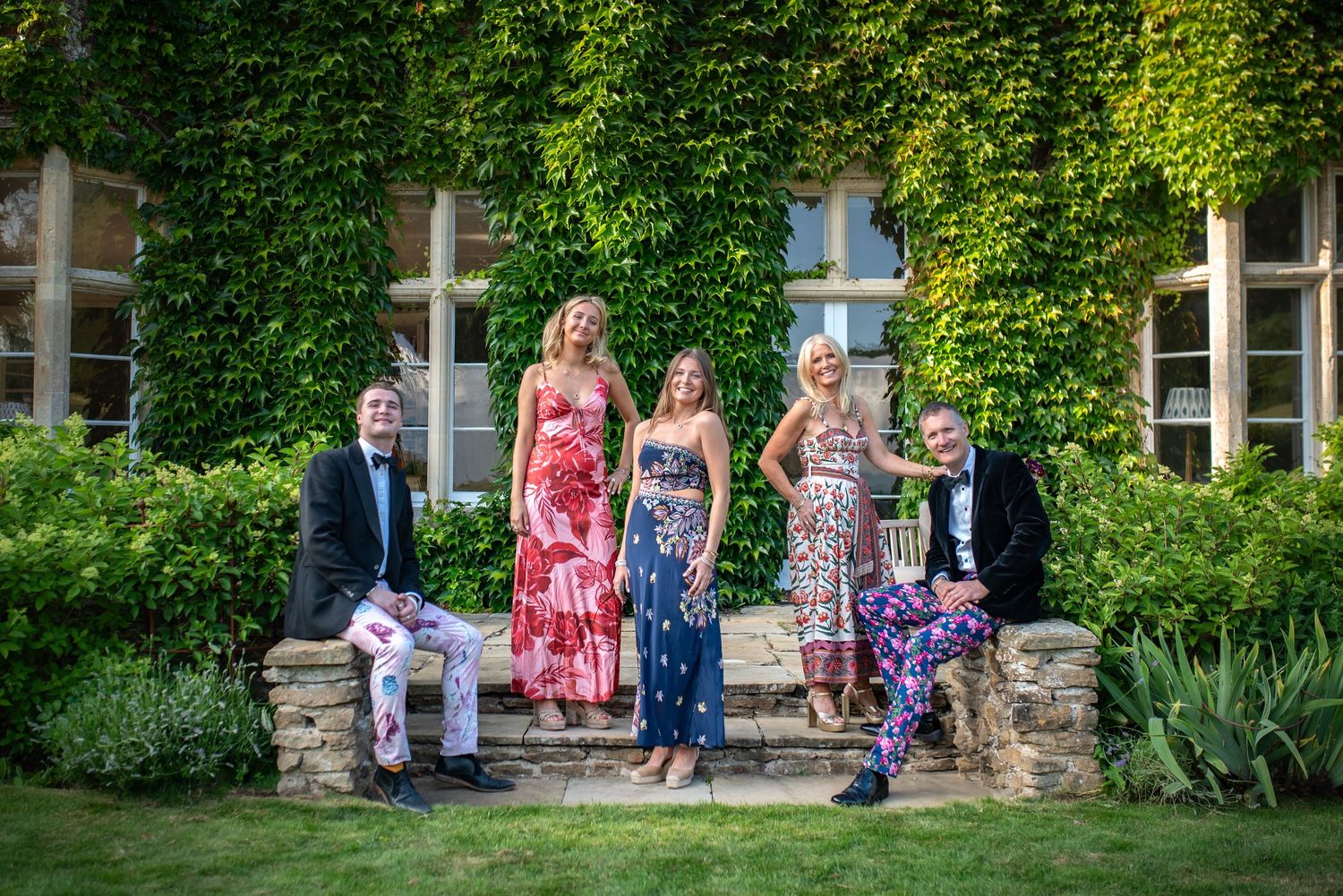Five elegantly dressed guests pose on stone steps outside a beautiful ivy-covered manor.