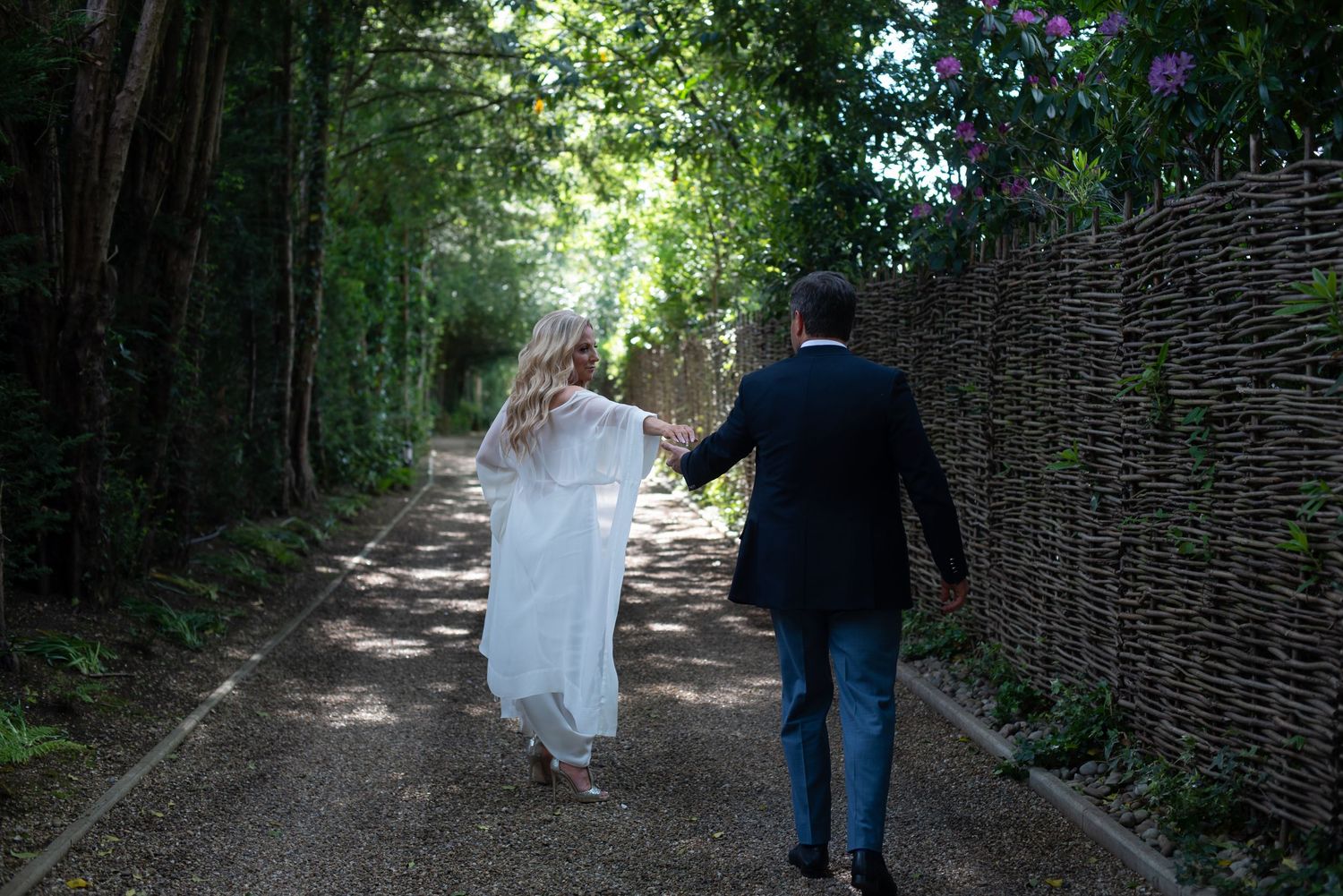 Bride and groom walk hand-in-hand down a tree-lined garden path on their wedding day.