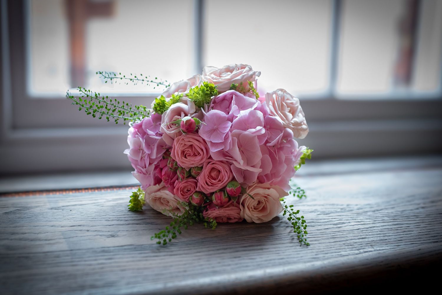 Bridal bouquet of pink hydrangeas and roses resting on a wooden windowsill in soft natural light.