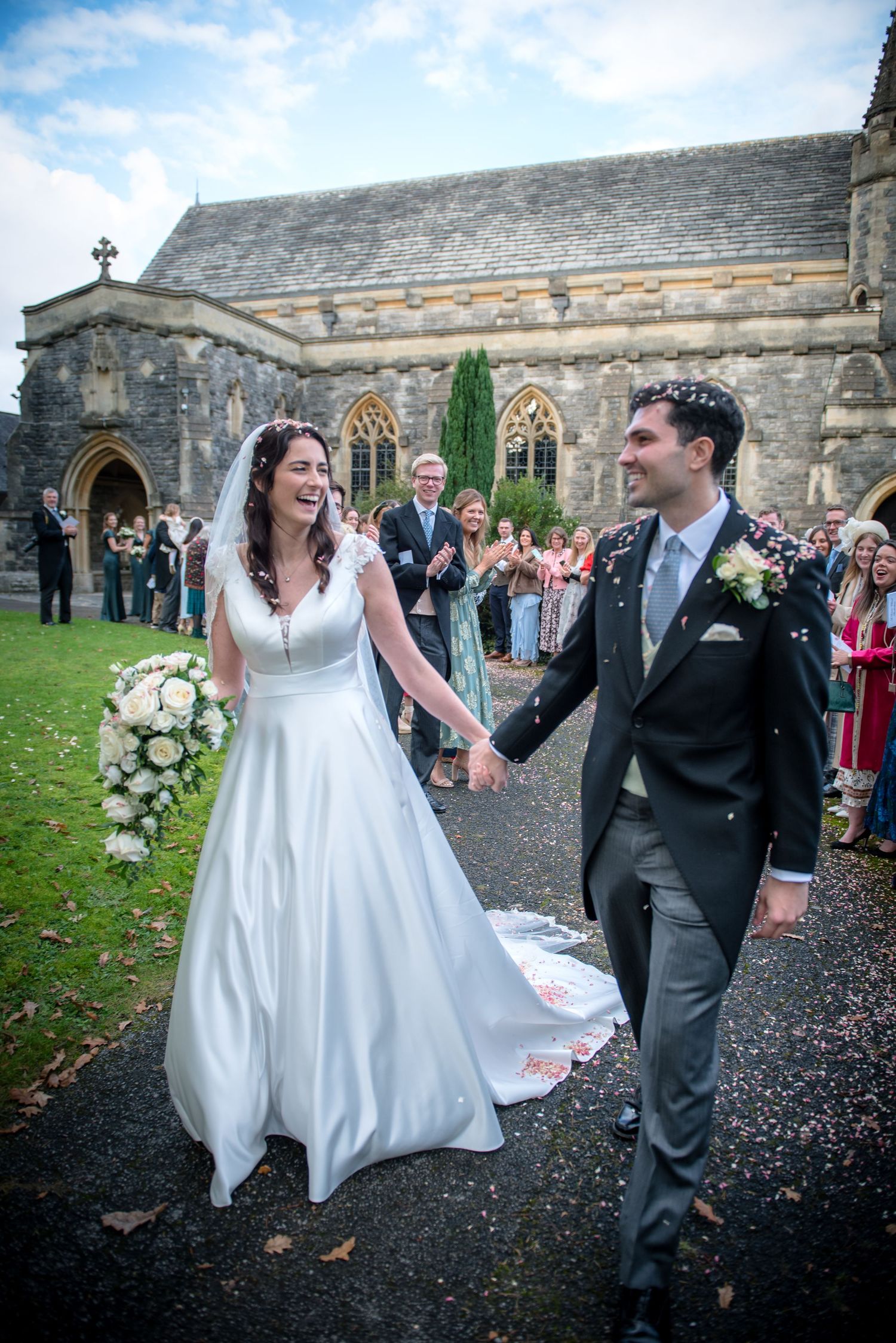 Joyful bride and groom walk hand in hand through confetti shower outside historic stone church. St Saviours Church, Brockenhusrt wedding