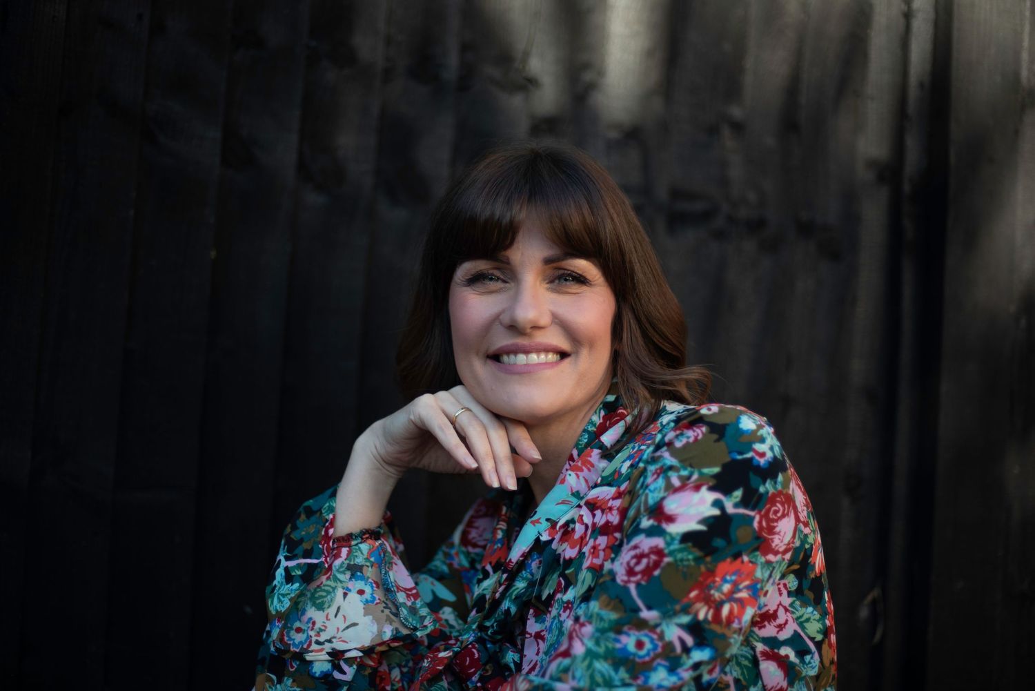 Smiling woman with dark hair poses against a dark textured background wearing a floral blouse.  business portrait