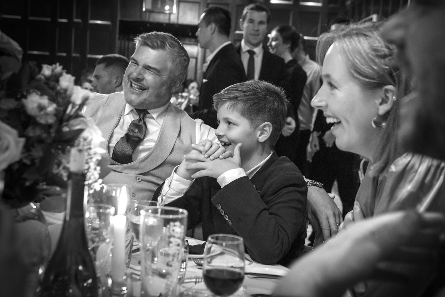 Black and white photo of wedding guests laughing and smiling at a candlelit wedding reception dinner table.