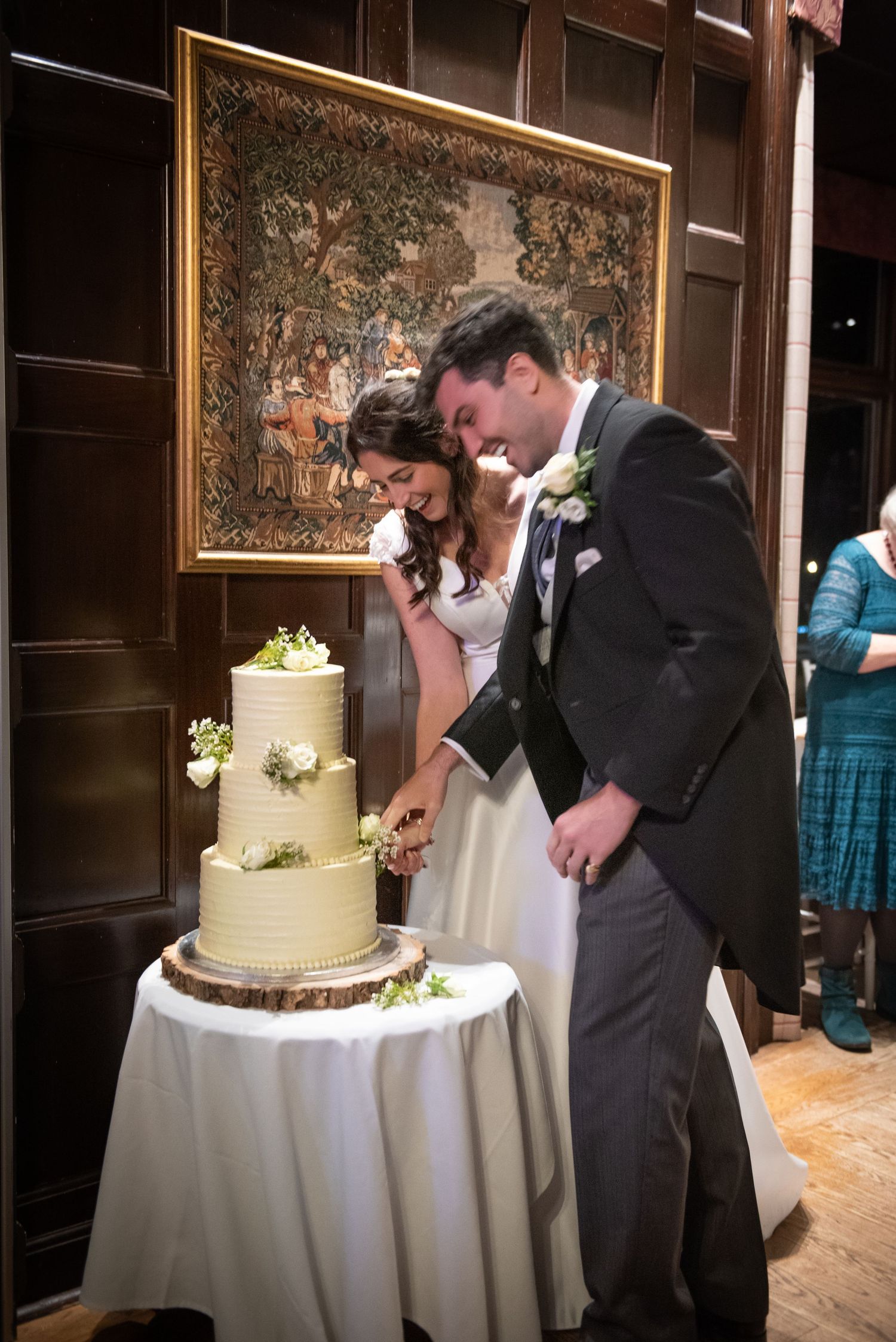 Bride and groom laughing together while cutting their three-tier white wedding cake at their reception.
