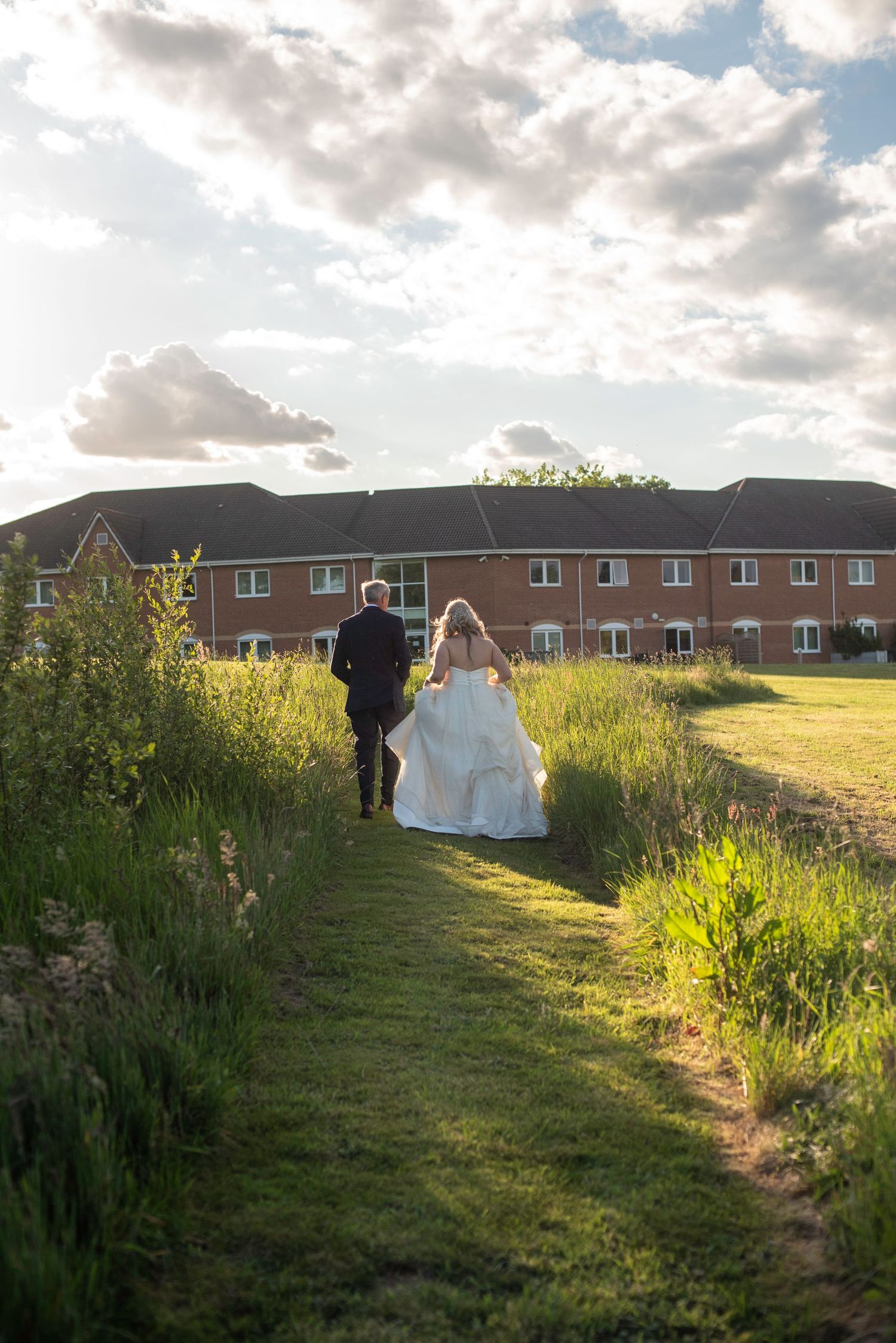 Bride and groom walking hand in hand through a lush green meadow toward a brick venue.