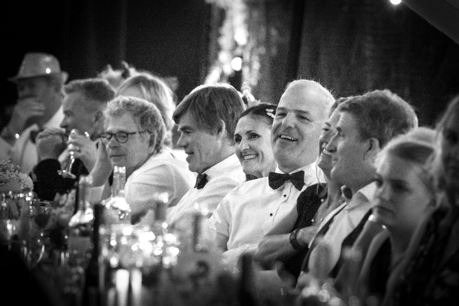 Black and white photo of wedding guests laughing and enjoying speeches at a formal reception dinner.