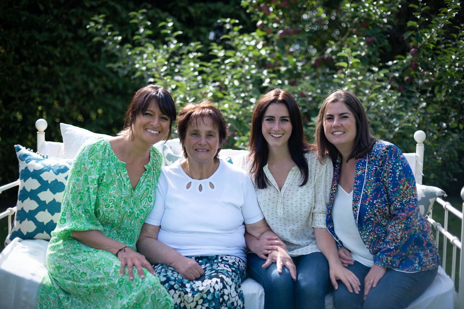 Four generations of women smiling together on a white bench in a garden setting.