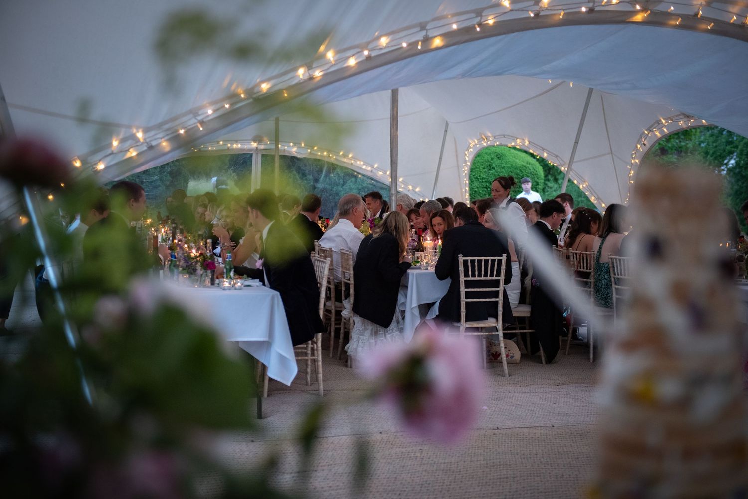 Guests dining under a beautifully lit wedding marquee adorned with fairy lights at dusk.
