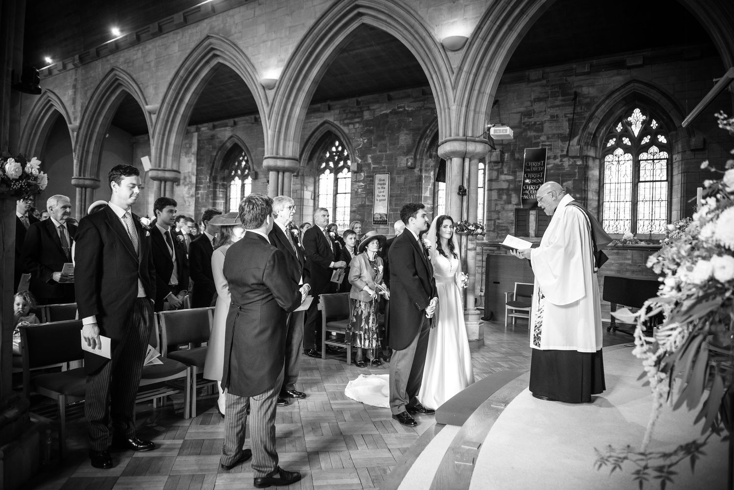 Wedding ceremony in a stone church with guests seated in pews as couple stands before officiant at the altar.  St Saviours Church