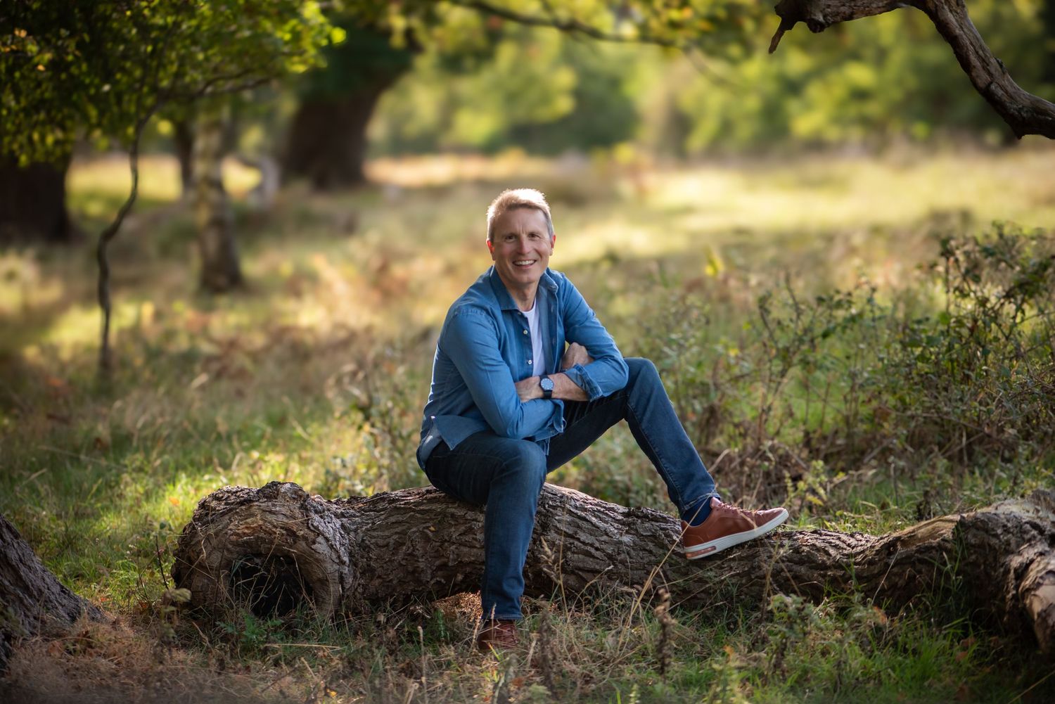 Man sitting on fallen log in sunny woodland park smiling at camera in casual blue outfit.