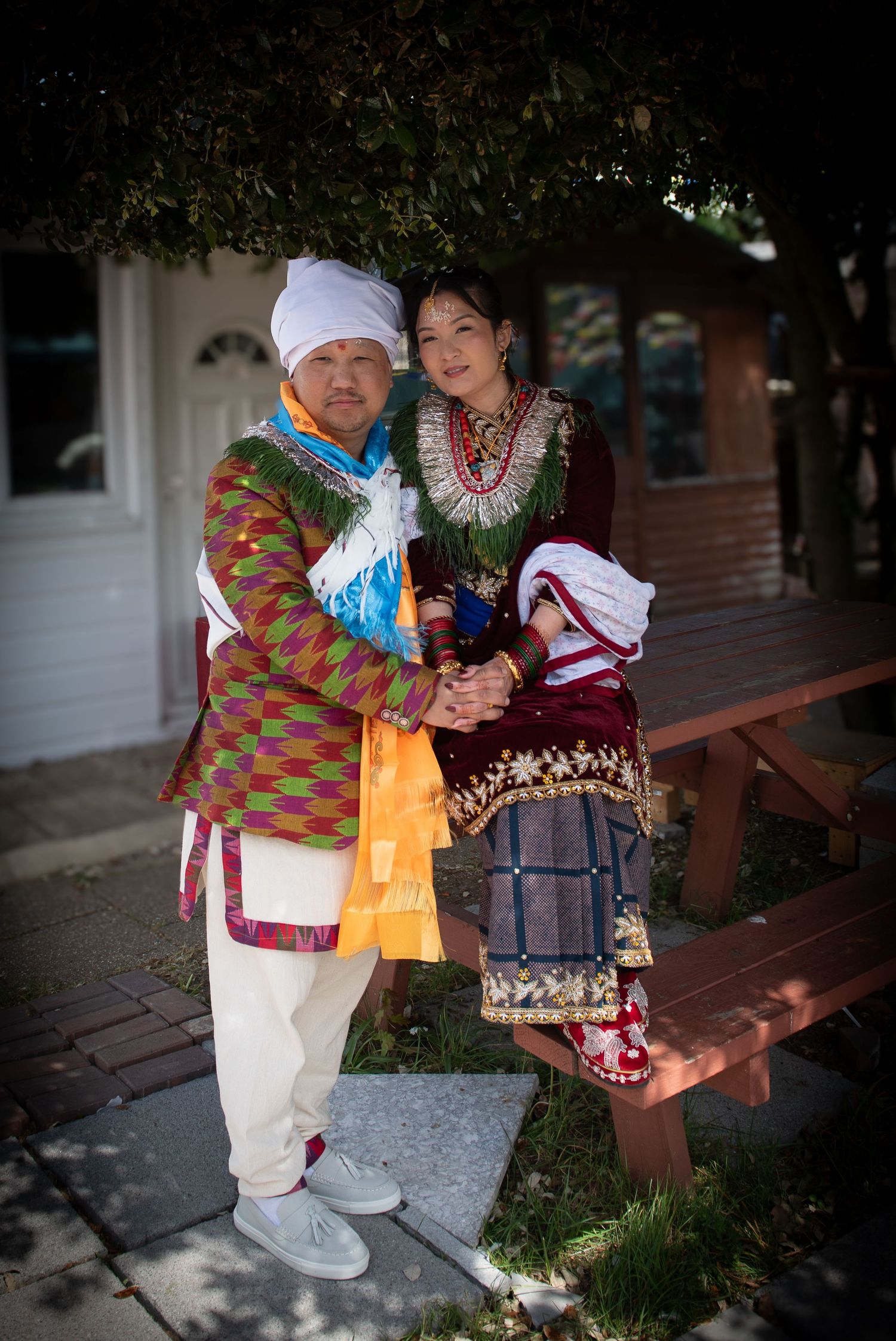 Couple in vibrant traditional cultural wedding attire embrace outdoors under a tree.