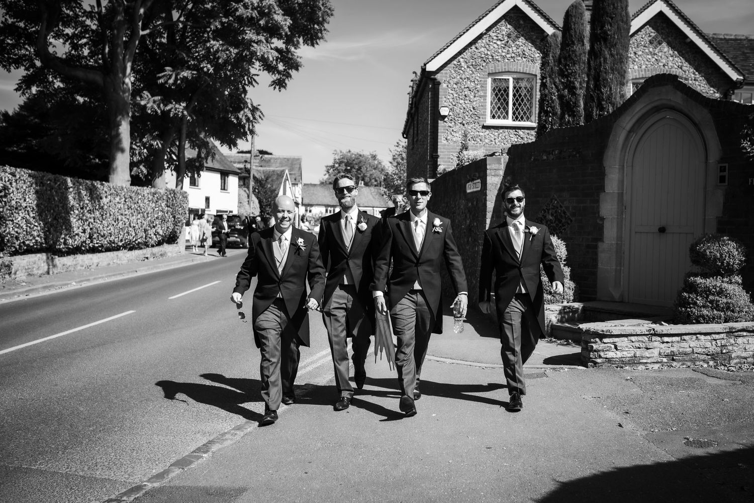Three groomsmen in dark suits walk confidently down a village road in a black and white photo.  St Michaels Church, Bray
