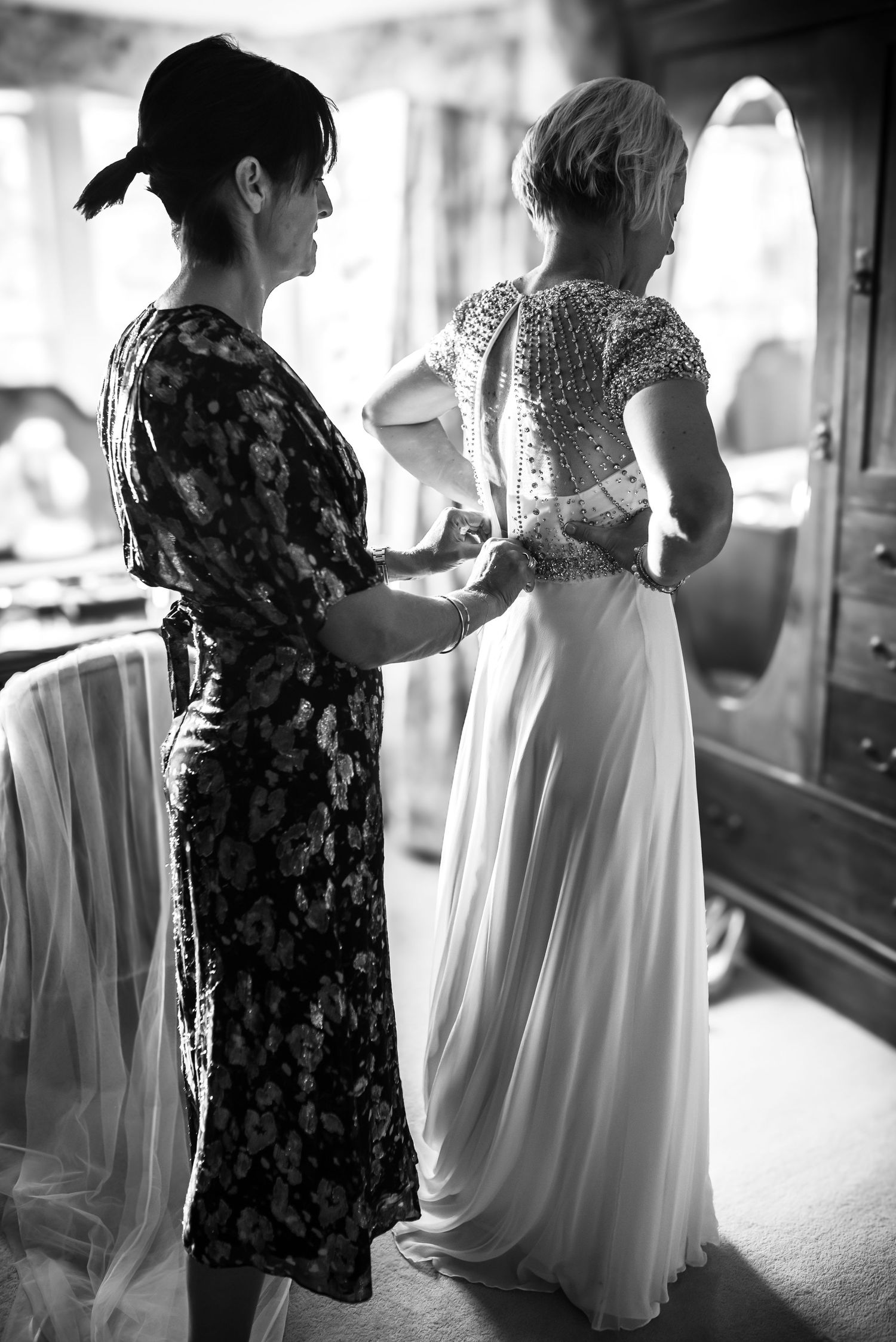 Black and white photo of mother helping bride fasten her beaded wedding dress before the ceremony.