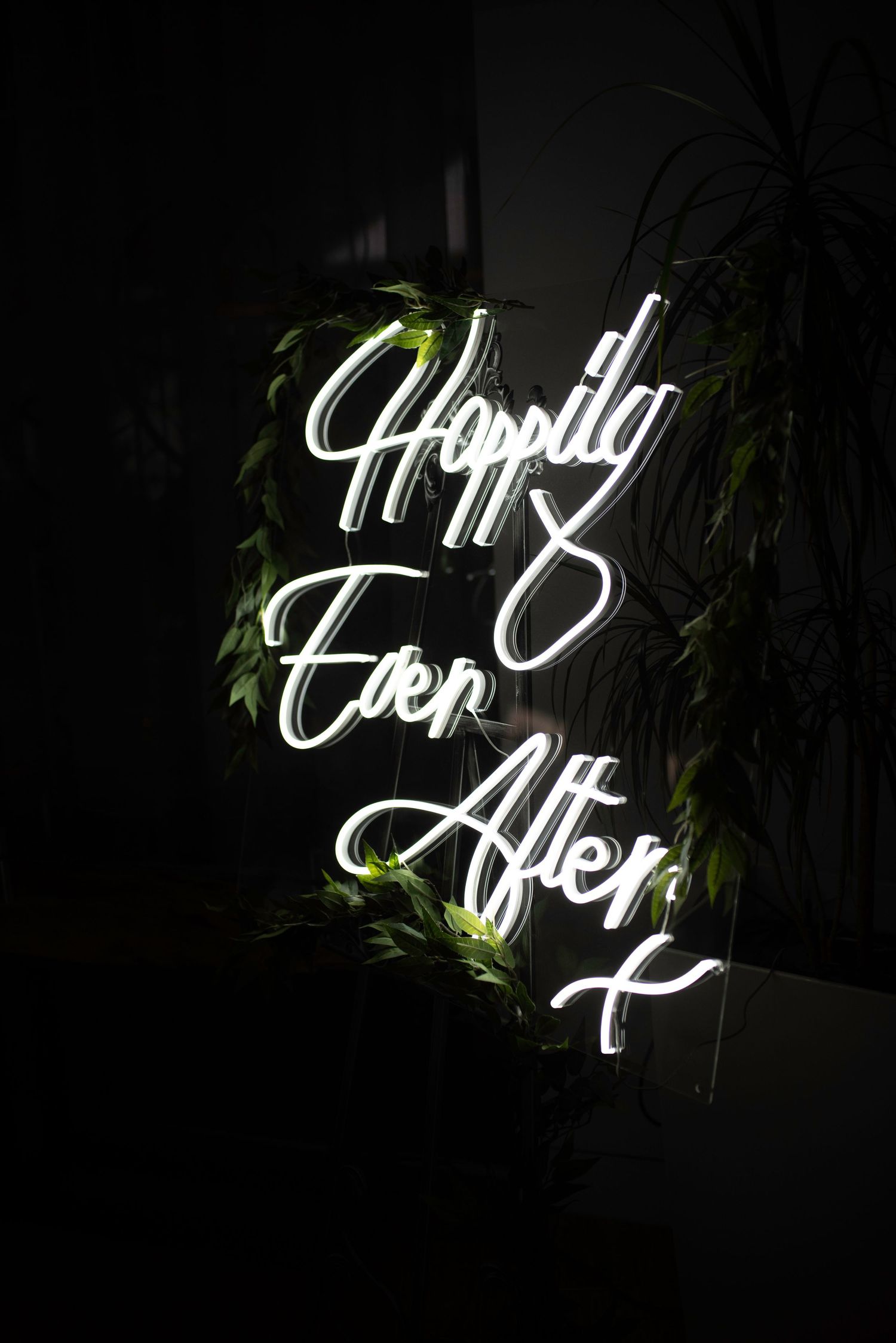 Neon sign reading Happily Ever After surrounded by green foliage in dark setting.