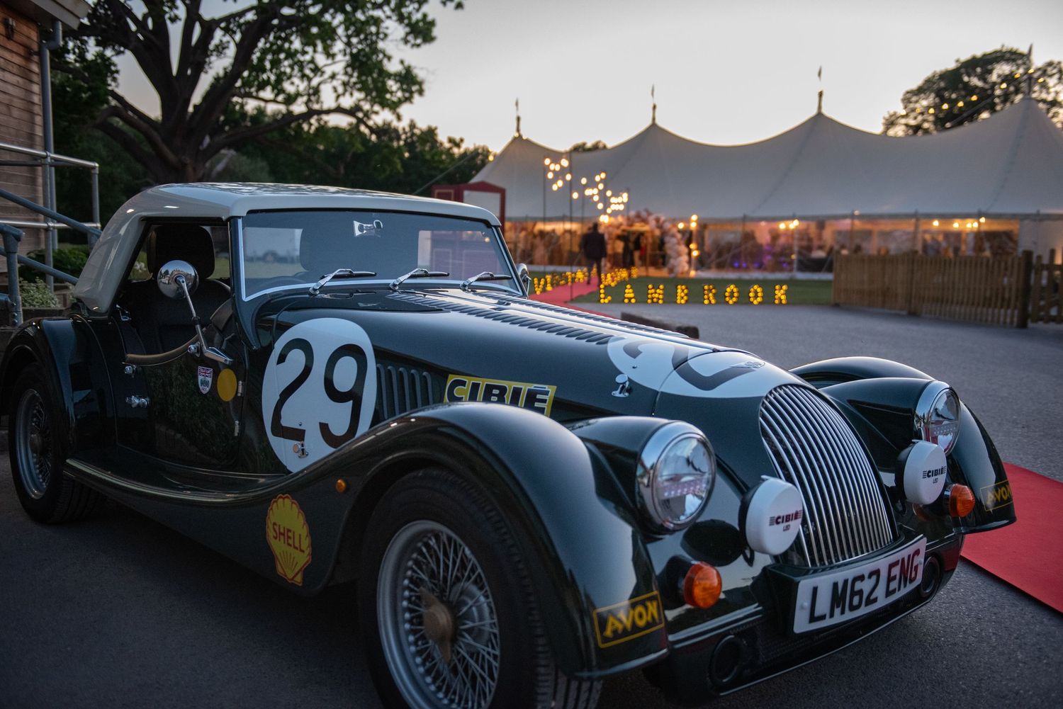 Vintage Morgan racing car number 29 parked outside illuminated marquee event venue at dusk.