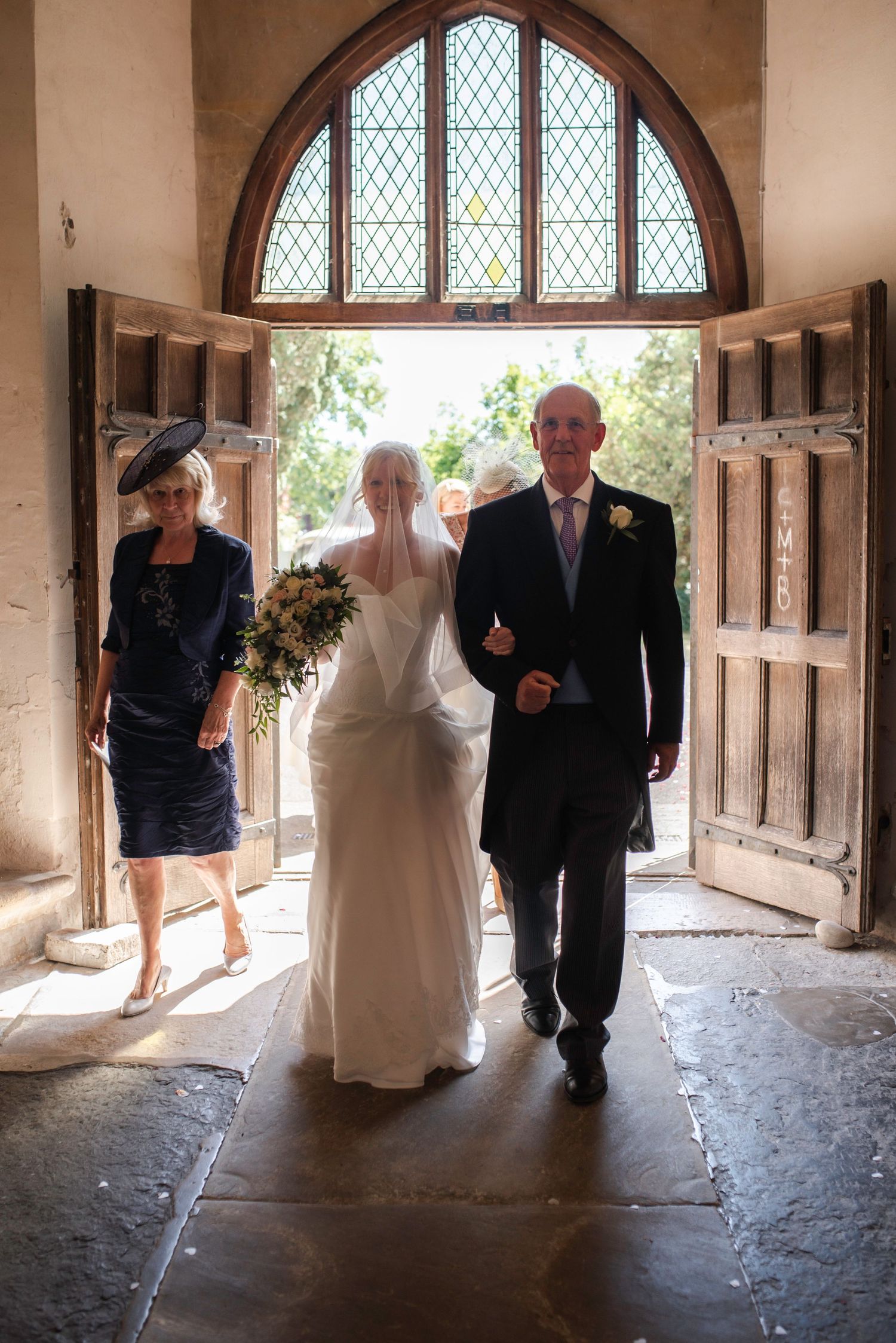 Bride in white gown walking down the aisle with her father through ornate church wooden doors. St Michaels Church, Bray