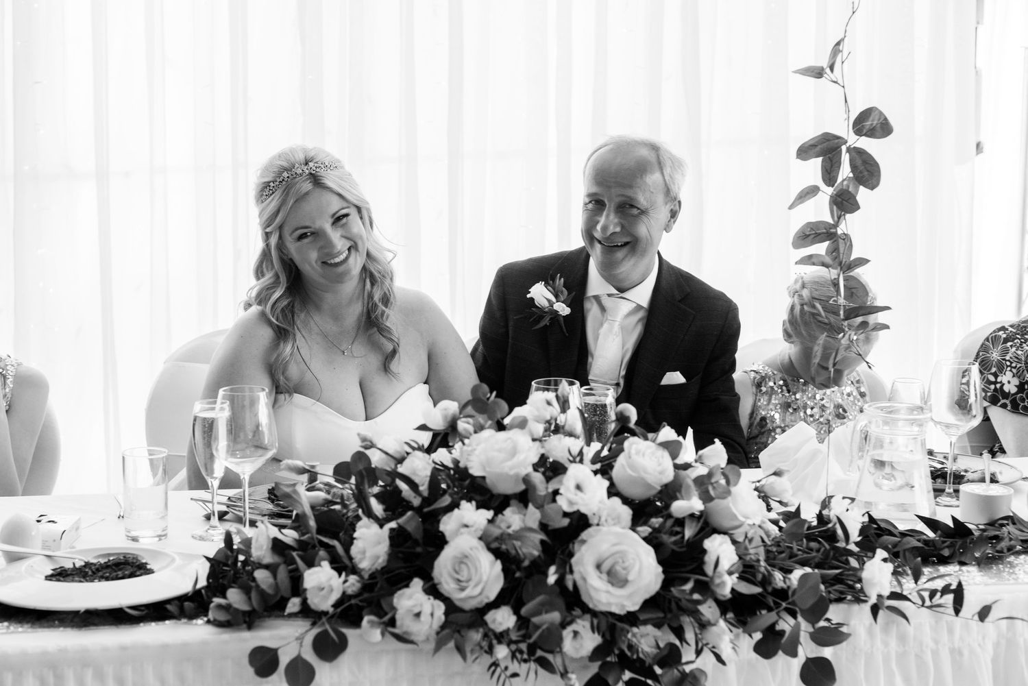 Smiling bride and groom seated at floral decorated head table during elegant wedding reception.