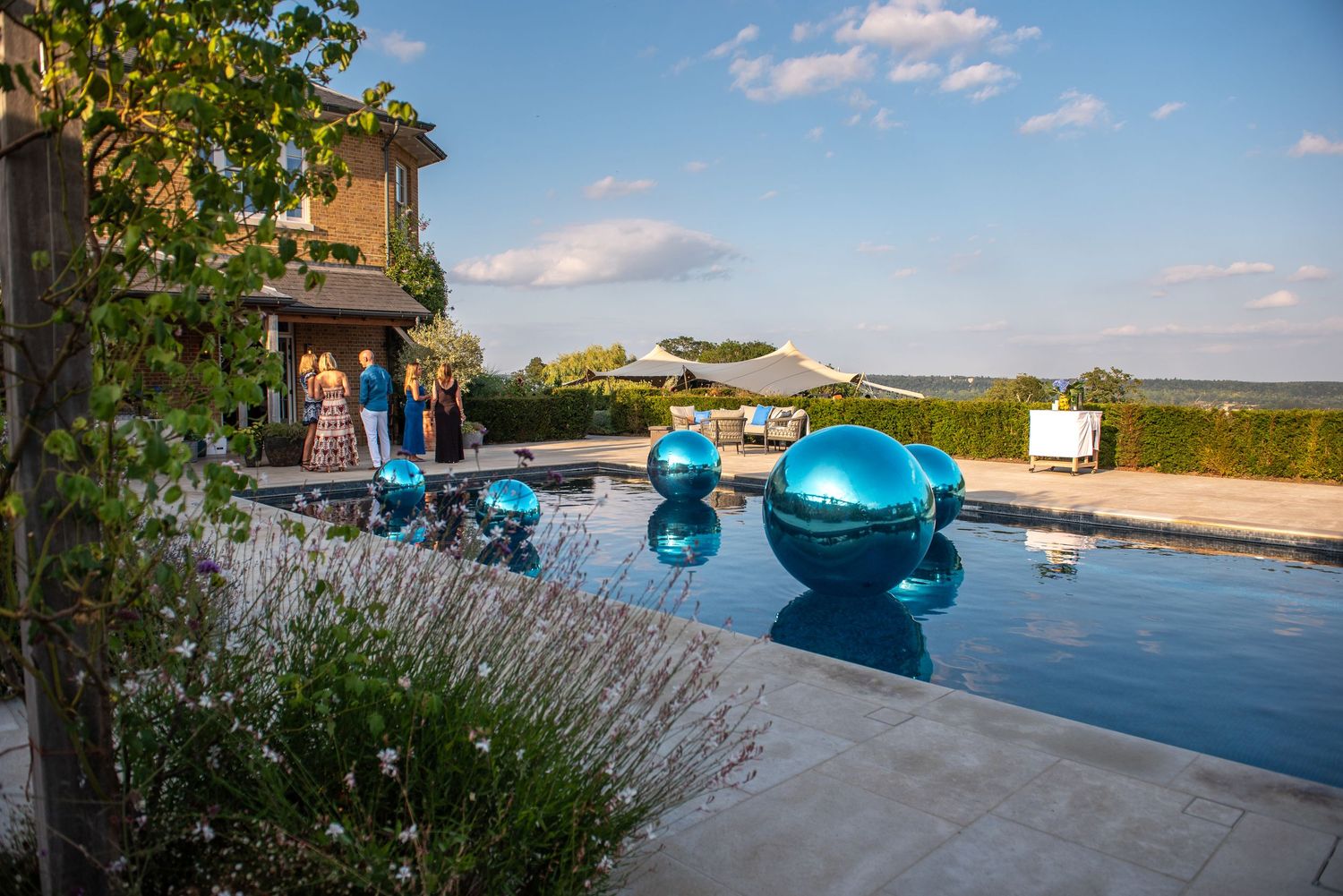 Elegant outdoor pool with blue reflective spheres and wedding guests at golden hour in countryside venue.