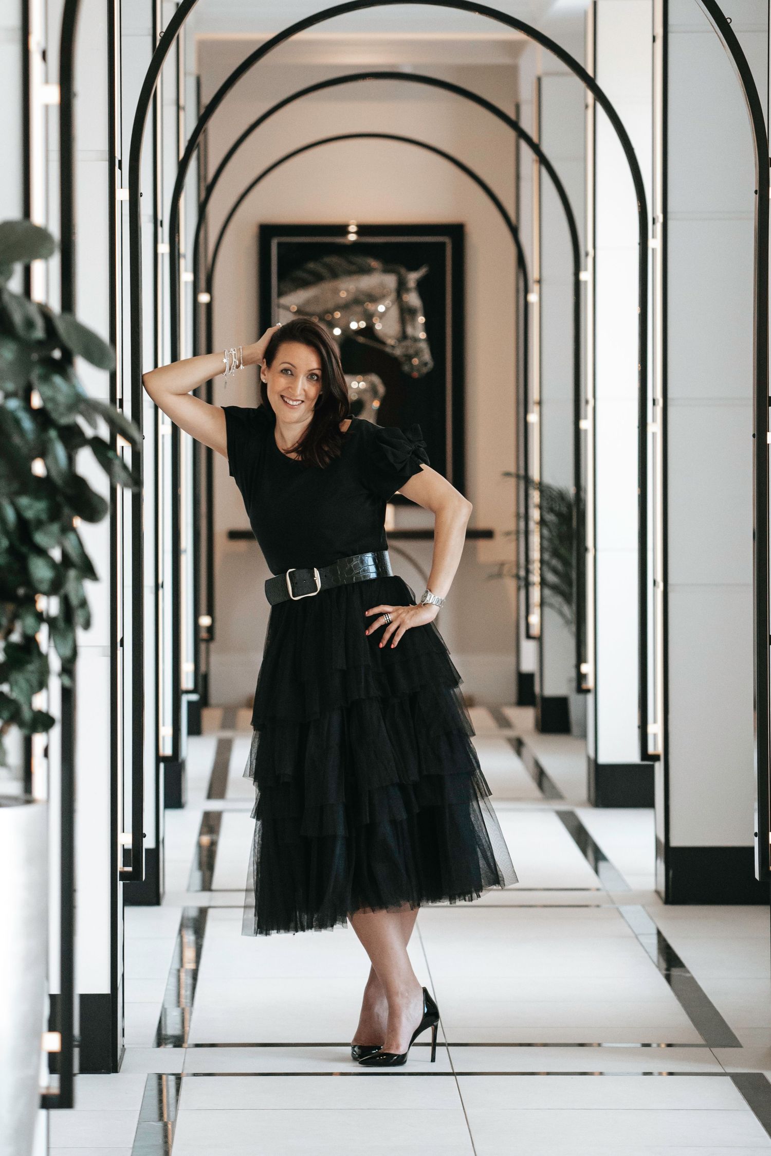 Smiling woman in black tulle skirt and belt poses in a luxury hotel corridor with arched mirrors.  business portrait
