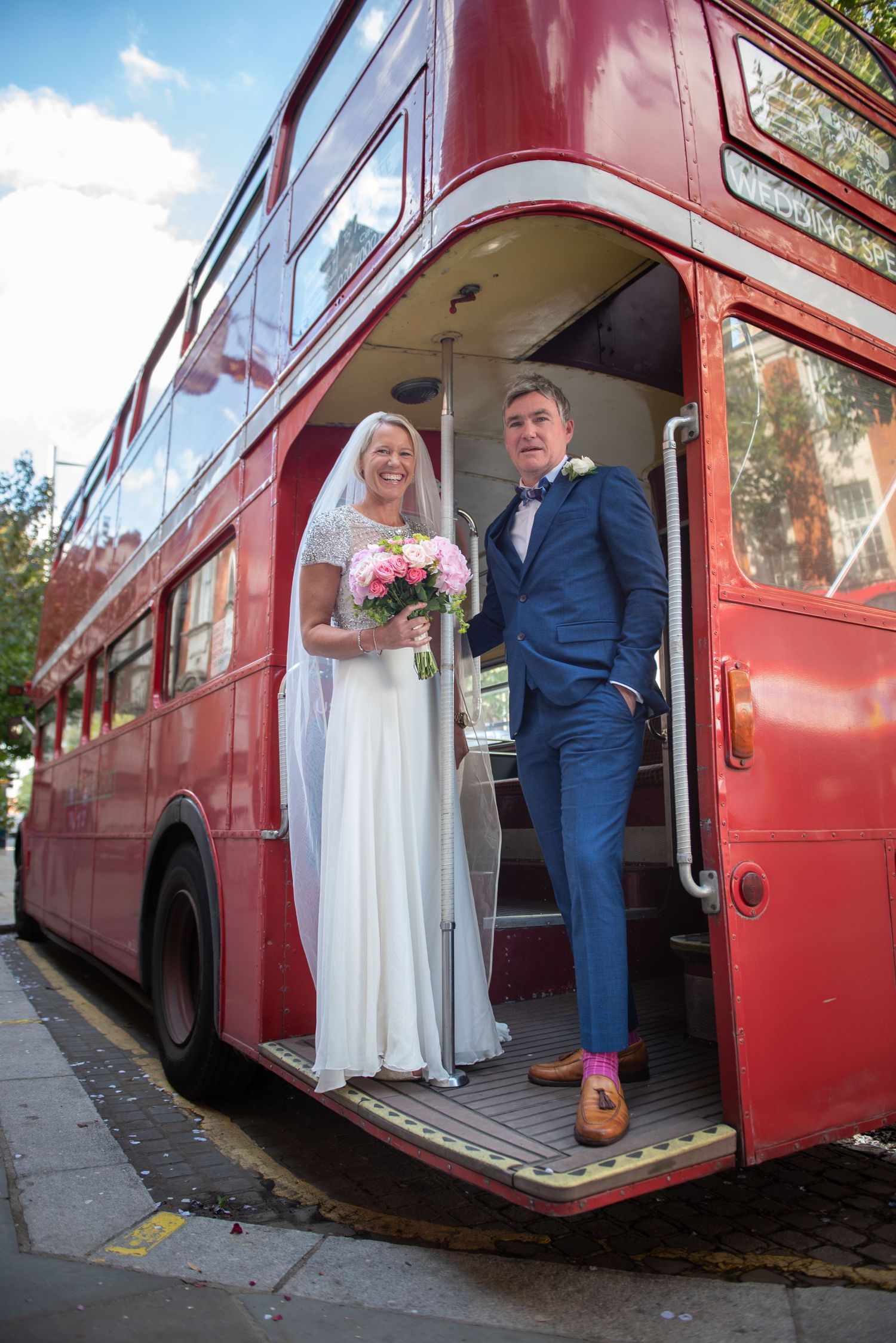 Bride and groom posing on steps of vintage red London double-decker wedding bus. Chelsea Town Hall Wedding