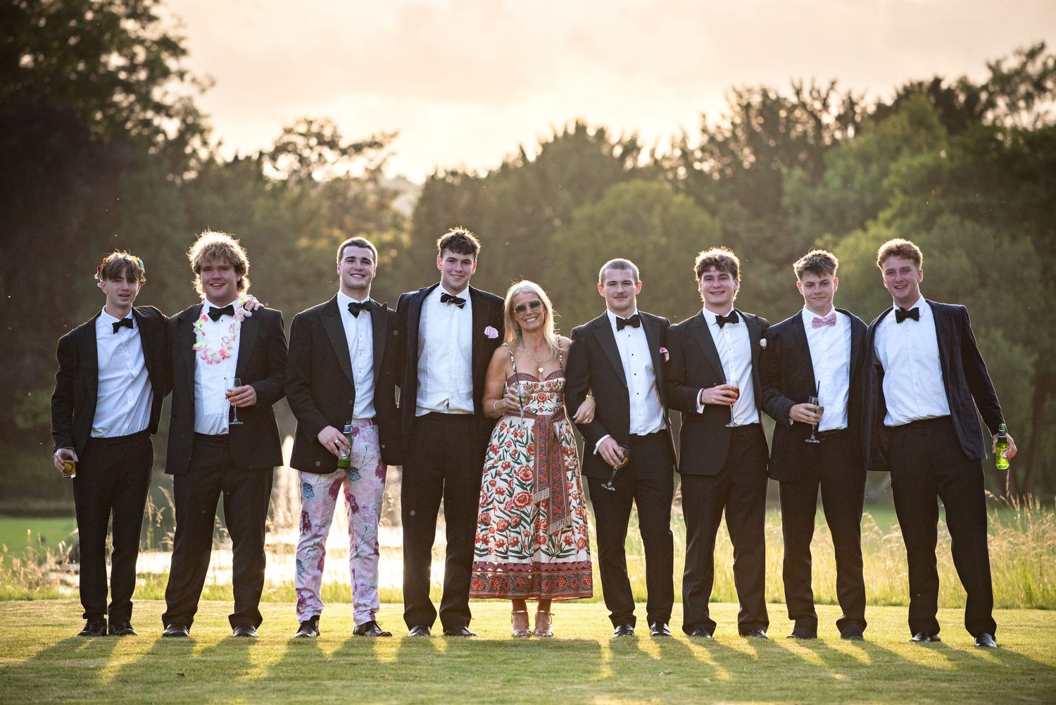 Group of young men in tuxedos with a woman in floral dress posing on green lawn at golden hour.