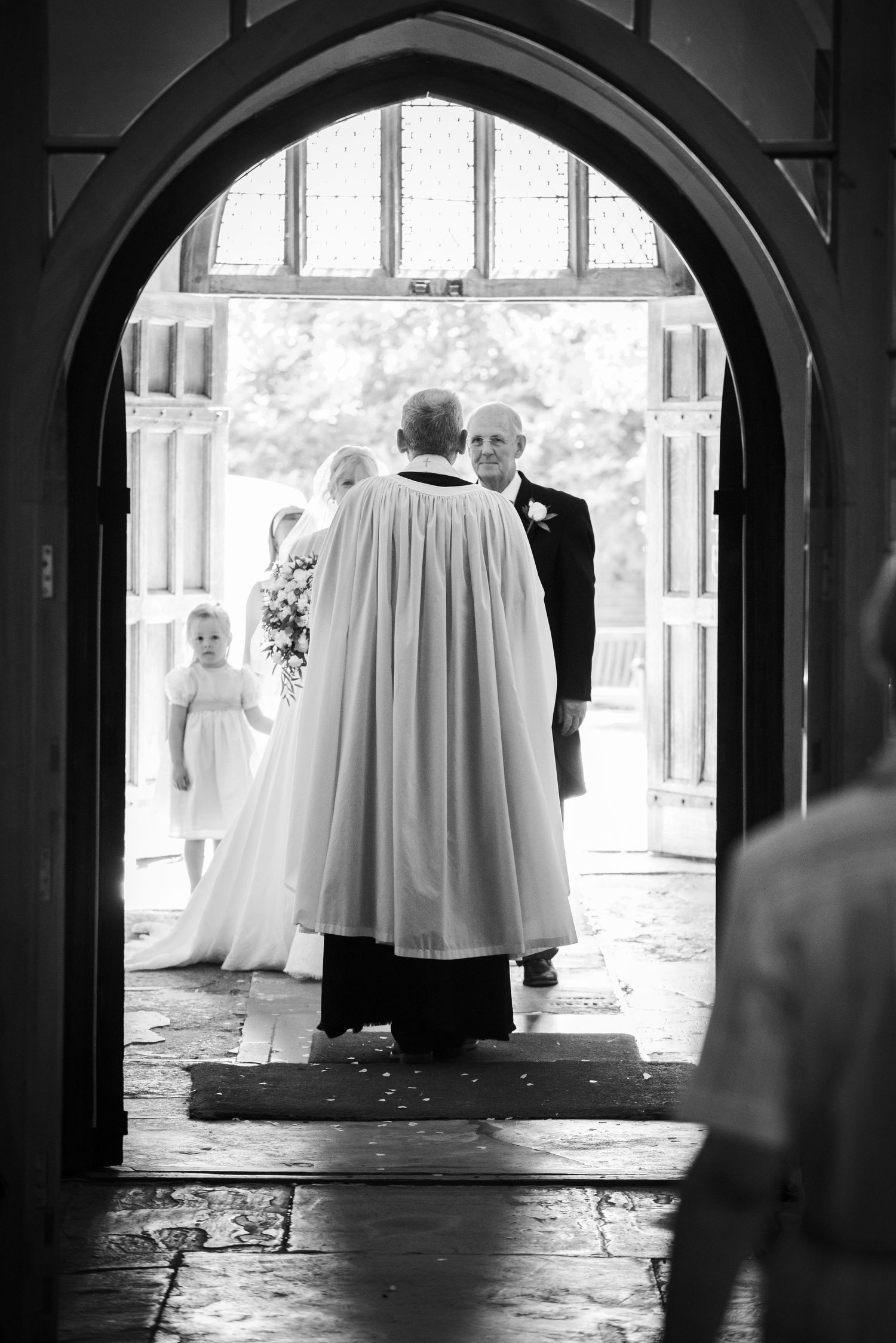 Priest greeting bride in white gown and flower girl at church entrance during wedding ceremony. St Michaels Church, Bray