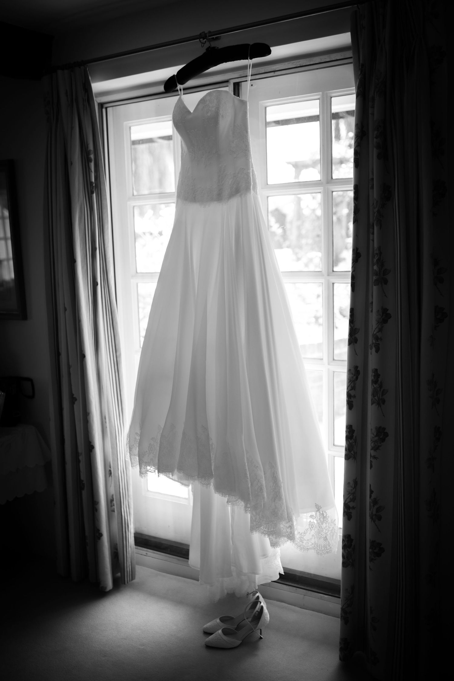 Elegant strapless wedding dress hanging in window with bridal heels below in black and white.