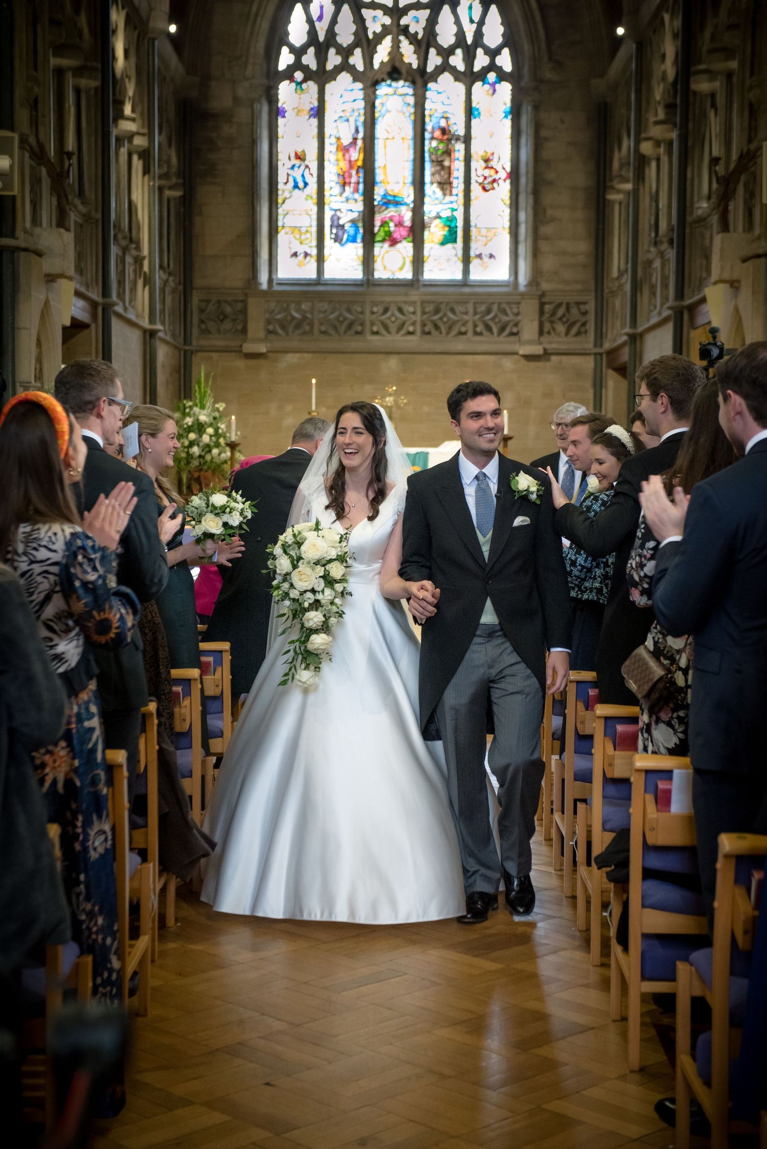 Smiling newlyweds walk down church aisle holding hands as guests clap with stained glass window behind. St Saviours Church Brockenhurst.