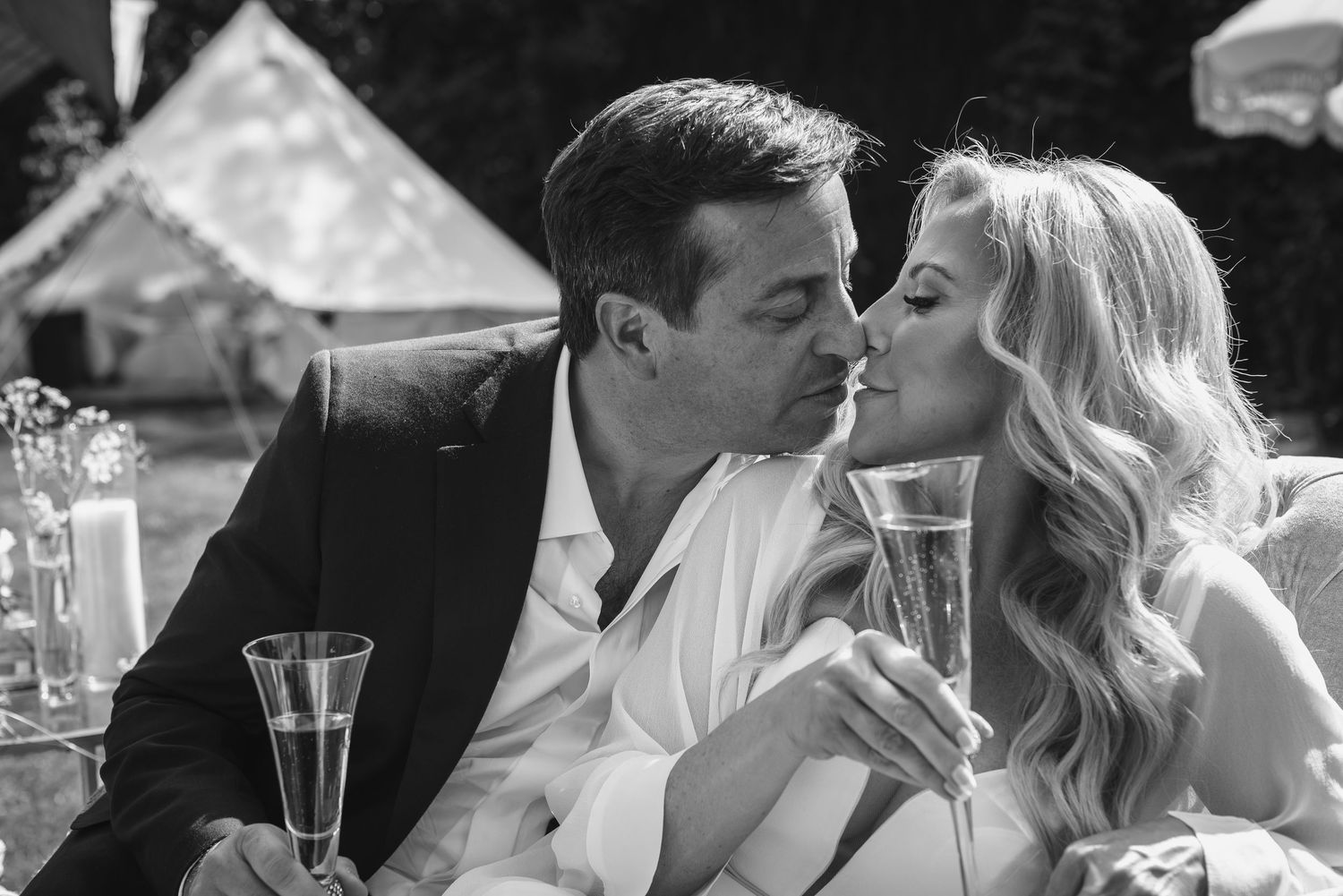 Black and white romantic photo of couple sharing a kiss while holding champagne flutes at outdoor celebration.