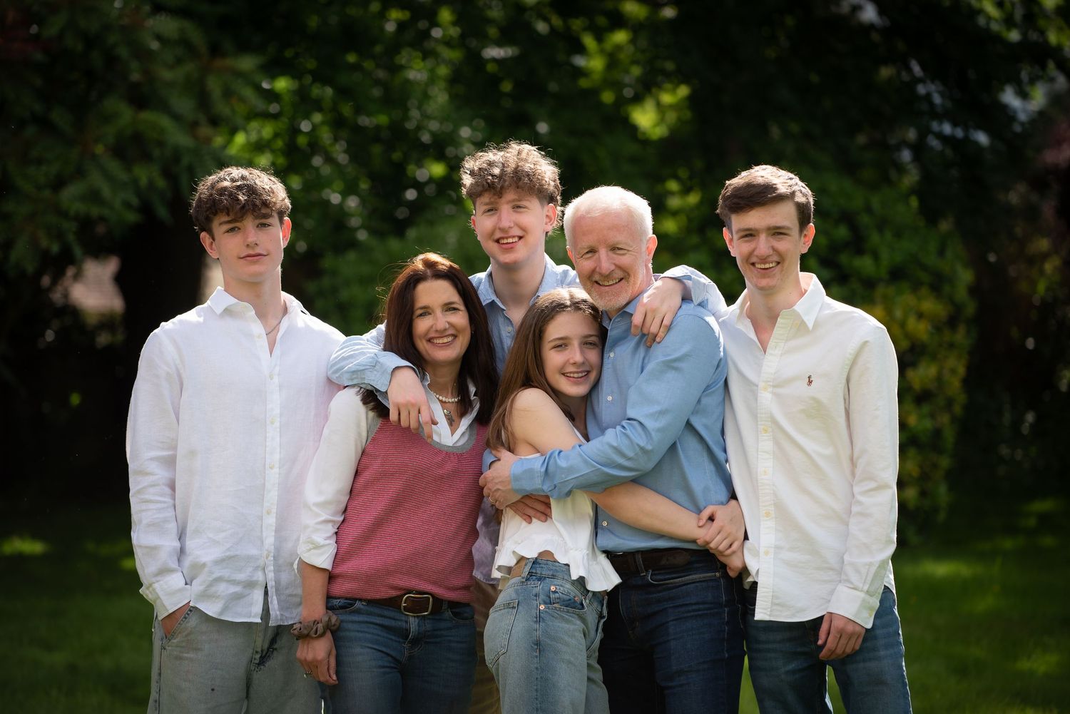 Happy family of six hugging and smiling together outdoors in a lush green garden setting.