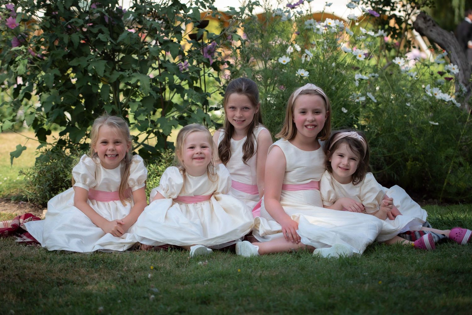Five young flower girls in white and pink dresses sitting together on grass in a garden.