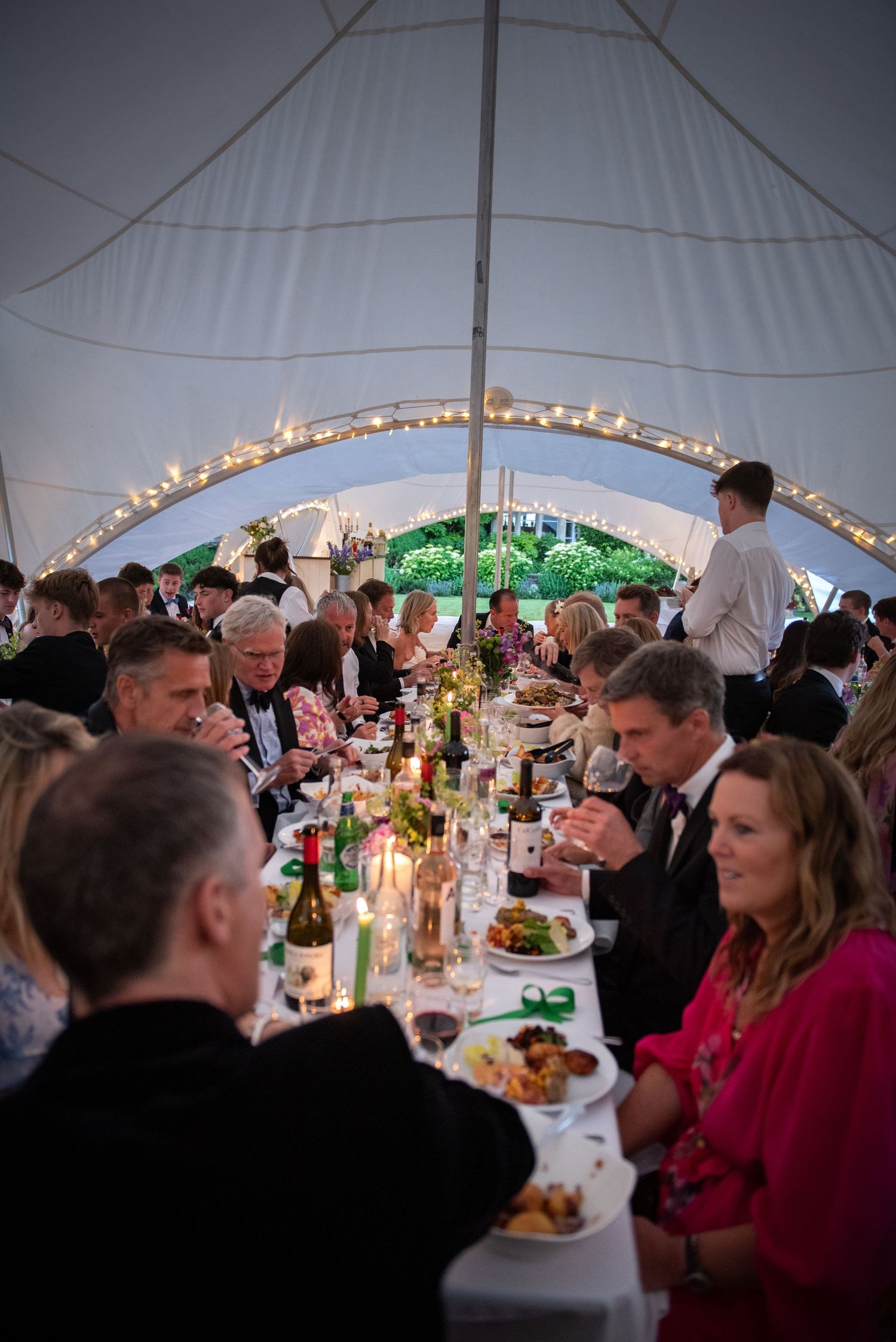 Elegant wedding reception dinner under a white tent with string lights, guests dining at long tables with candles.
