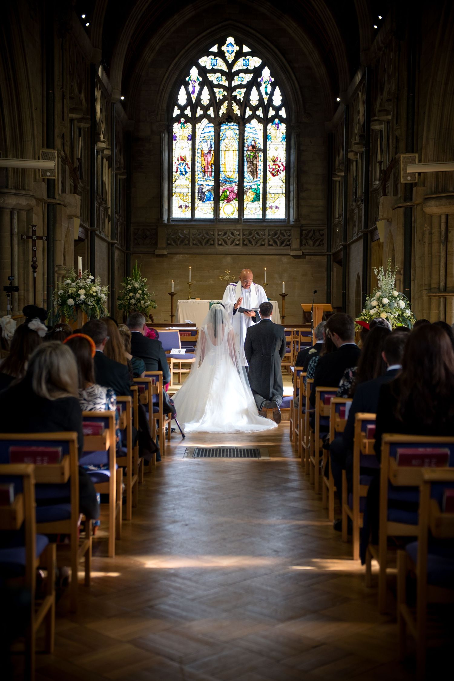 Bride and groom stand before officiant at church altar with colourful stained glass window in background. St Saviours church.
