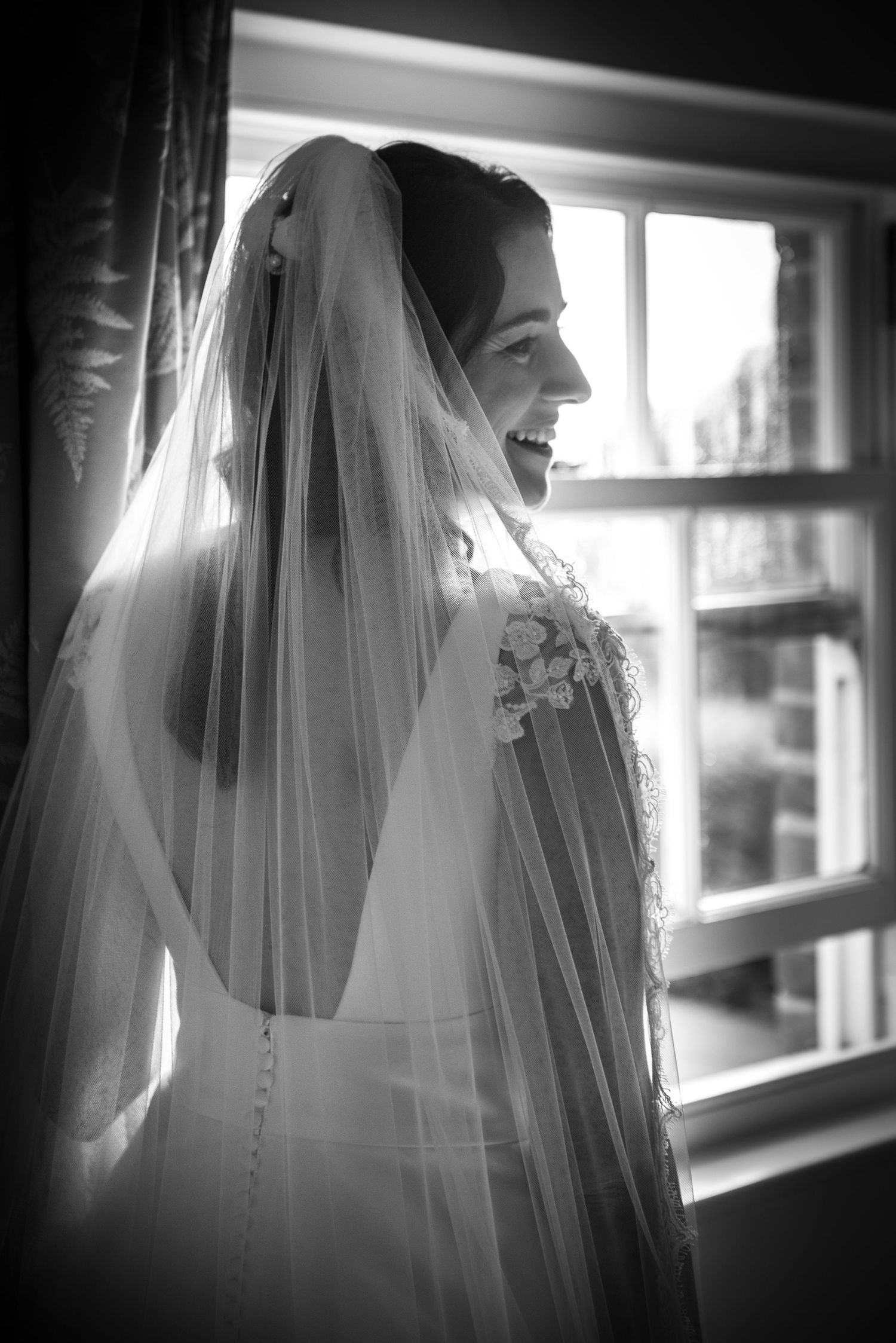 Radiant bride in veil smiling by a window in black and white portrait.