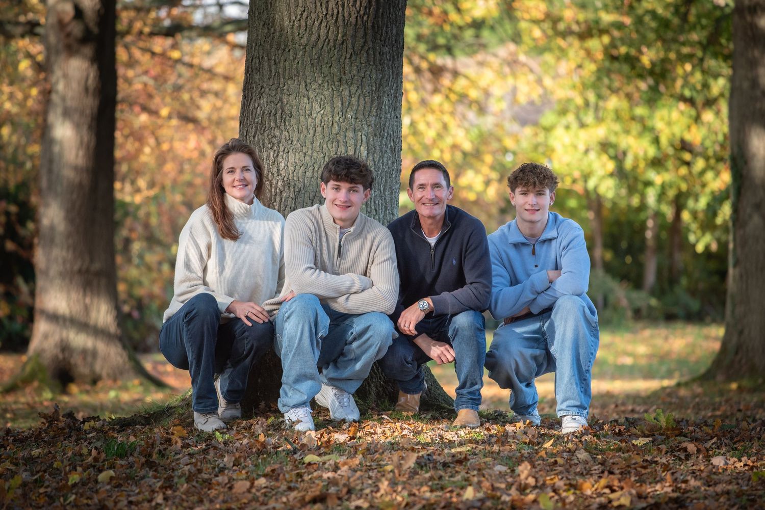 Family of four posing by a large tree trunk in a park with beautiful autumn foliage.