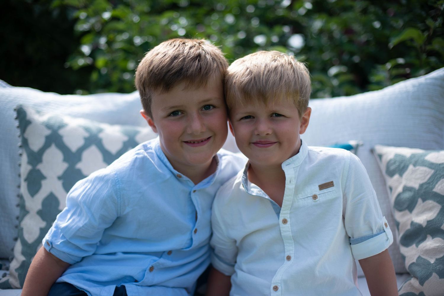 Two young brothers in light blue shirts smile together on a cushioned outdoor bench. family portrait