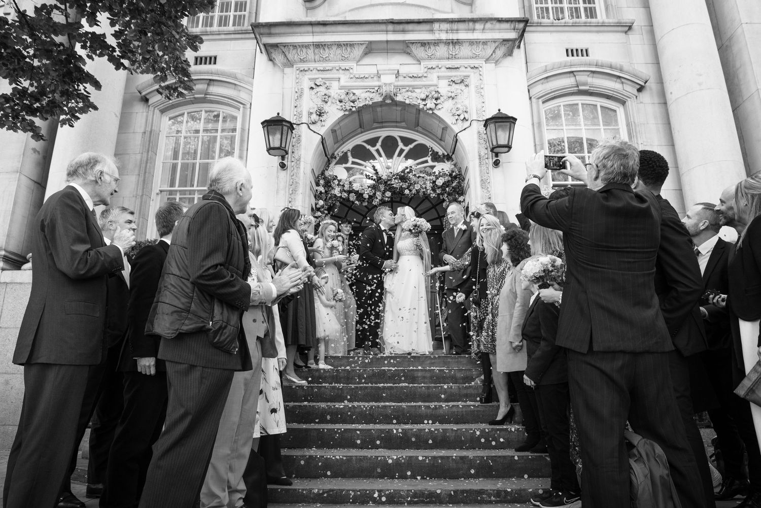 Black and white photo of wedding couple exiting grand building with guests throwing confetti on steps.  Chelsea Town Hall, London Wedding