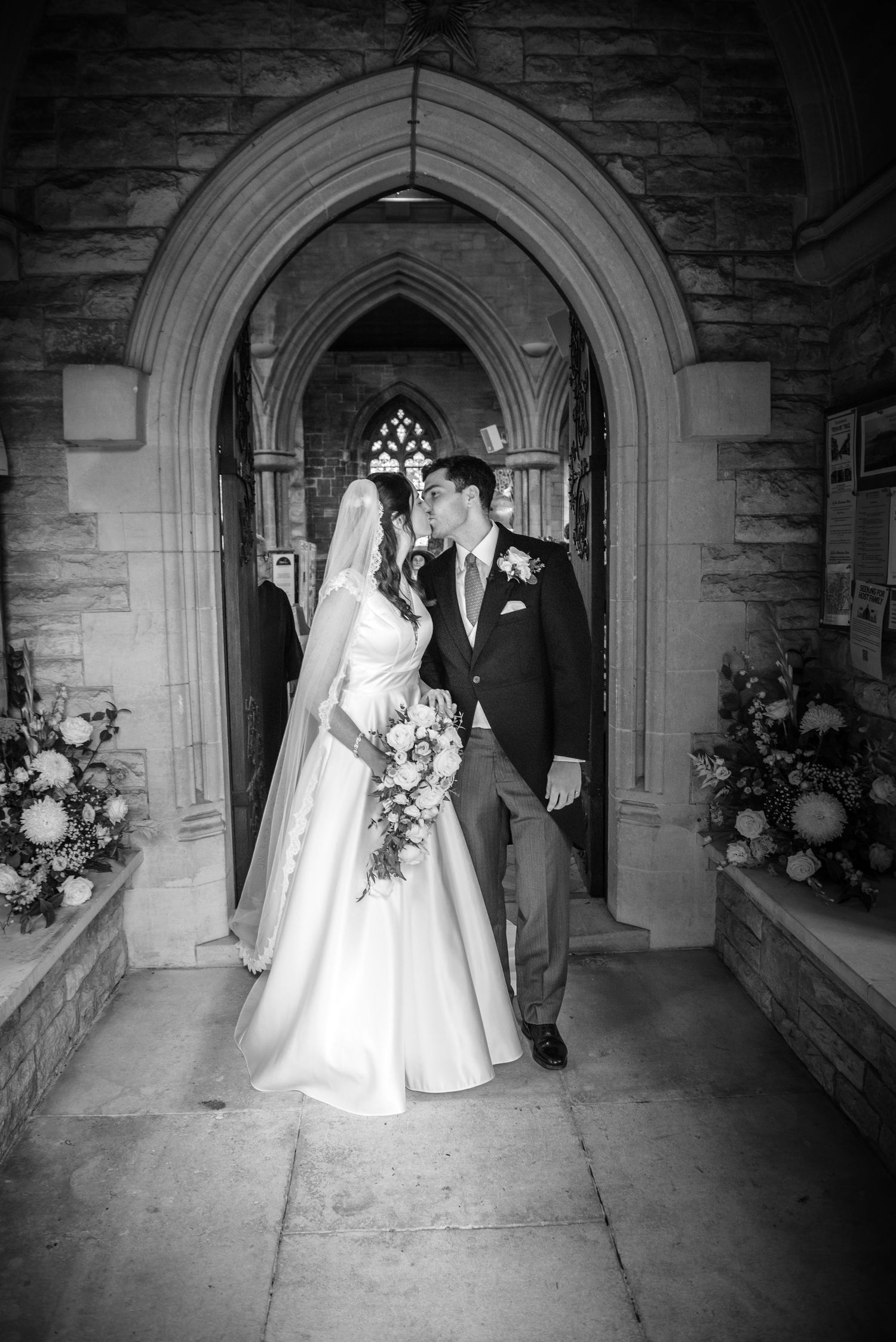 Black and white photo of a bride and groom sharing a kiss under a Gothic stone church archway.