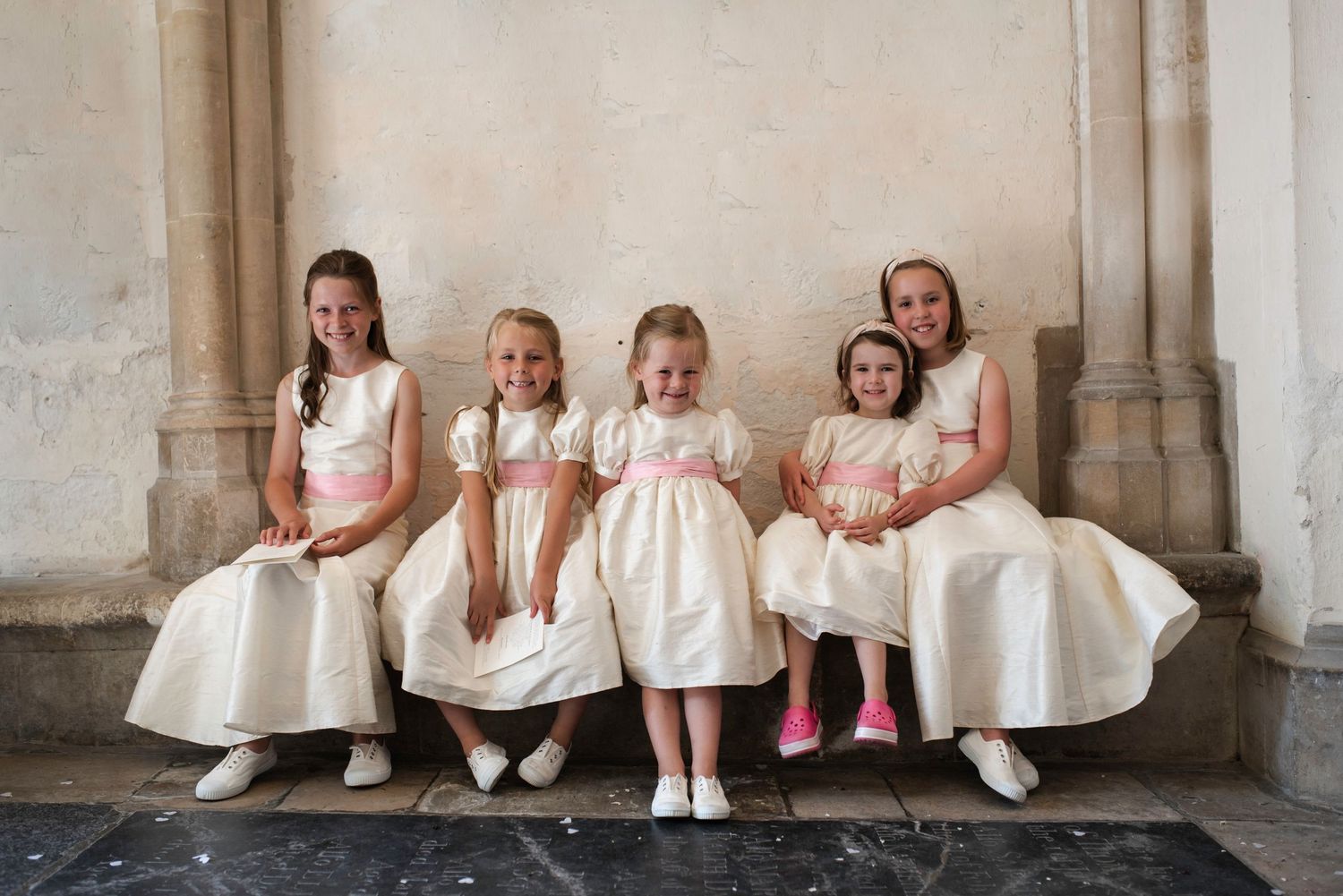 Five flower girls in matching ivory dresses with pink sashes sitting on a stone bench inside a church at St Michaels Church, Bray