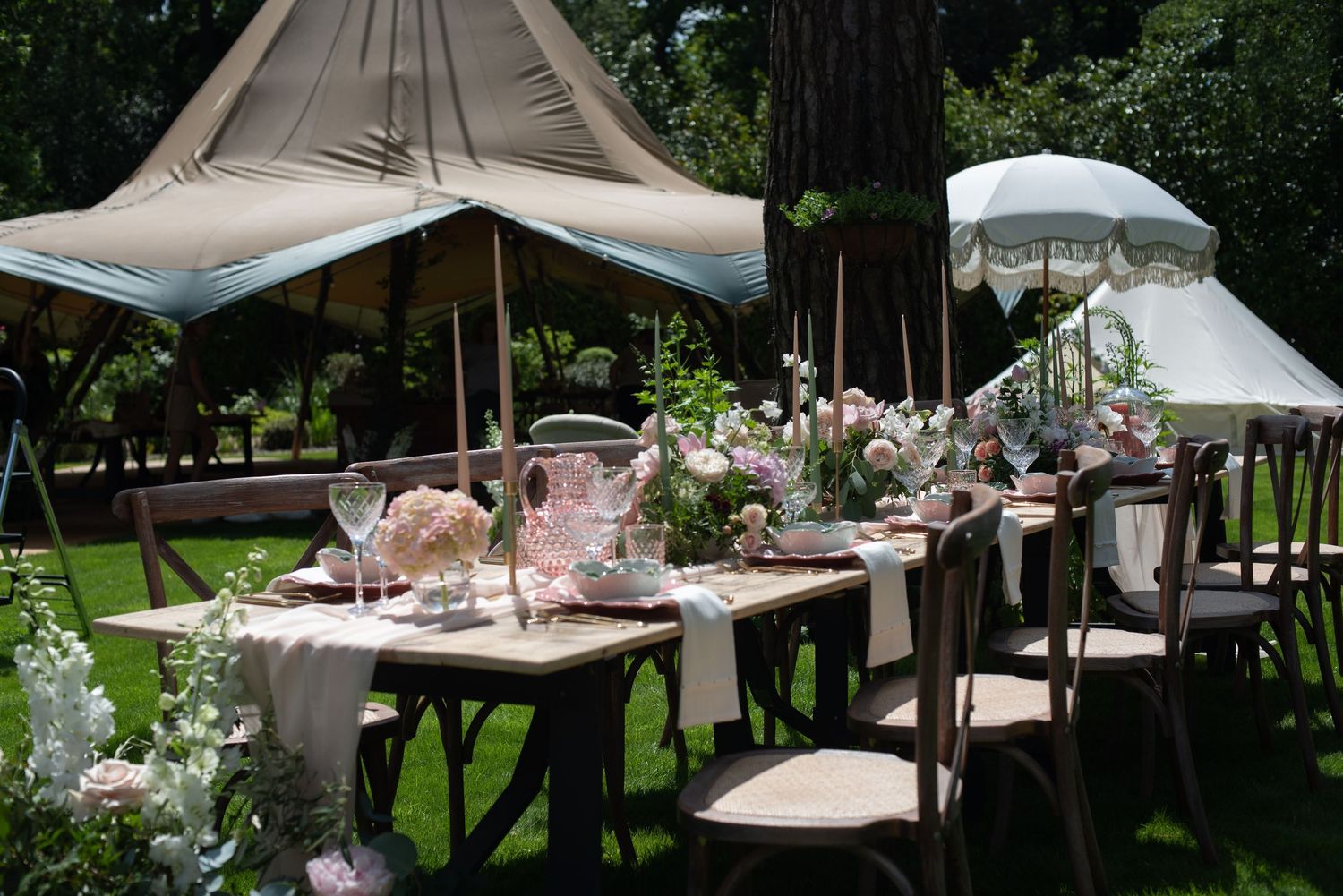 Romantic al fresco wedding table set under a canopy tent with pink blooms, cross-back chairs, and elegant tableware.