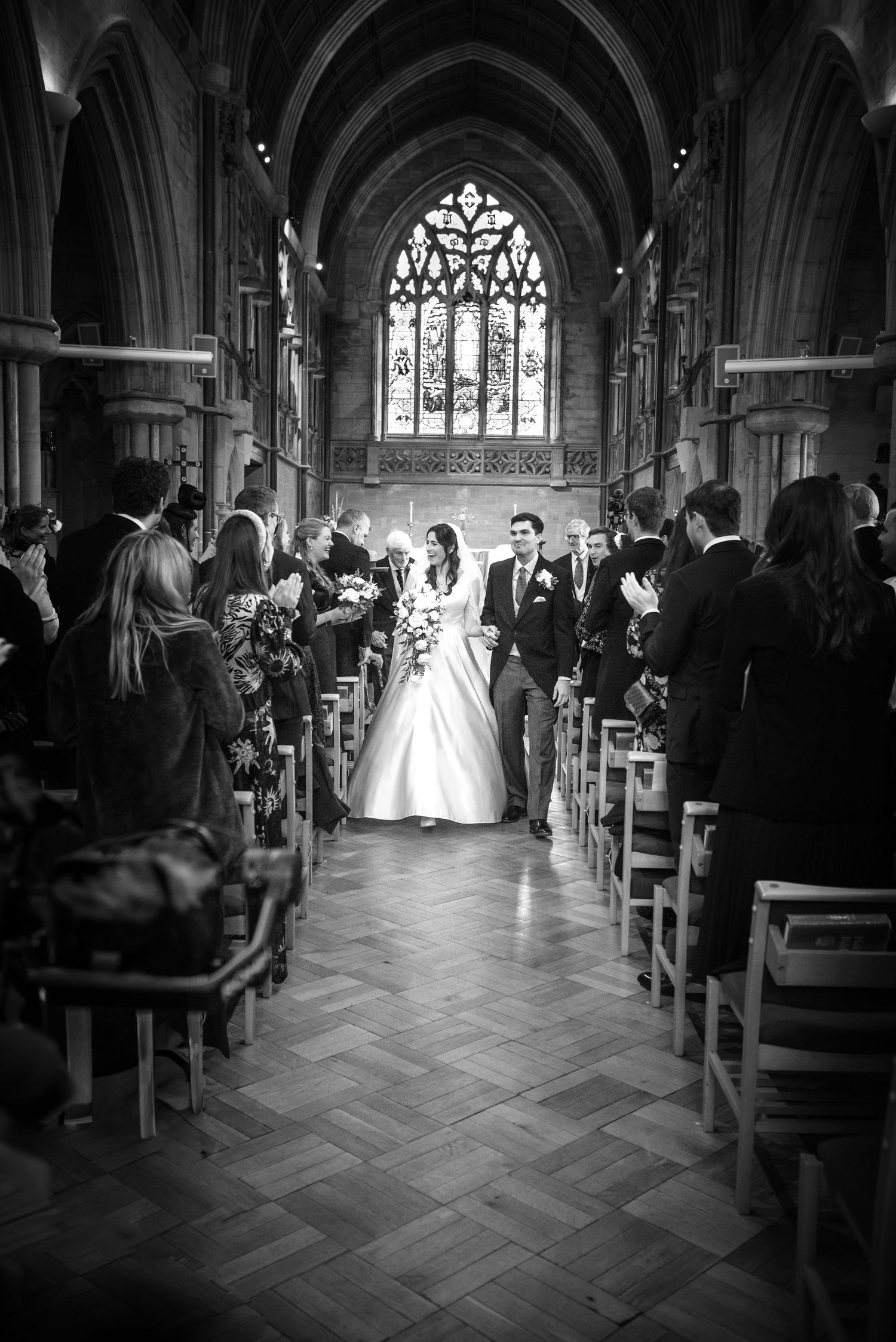 Black and white photo of newlyweds walking down church aisle with guests applauding on both sides. St Saviiours Church, Brockenhsurt wedding