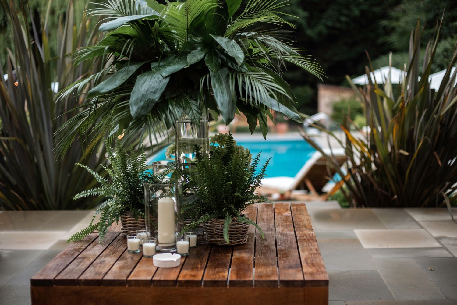 Elegant outdoor wedding table with tropical greenery, candles, and glass lanterns beside a pool.