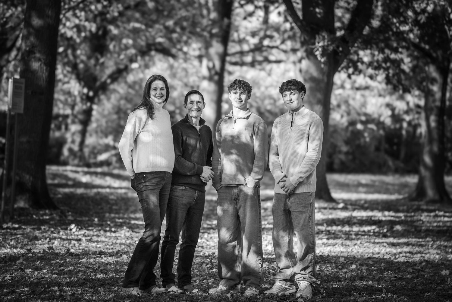 Black and white portrait of a family of four standing together in an autumn woodland.