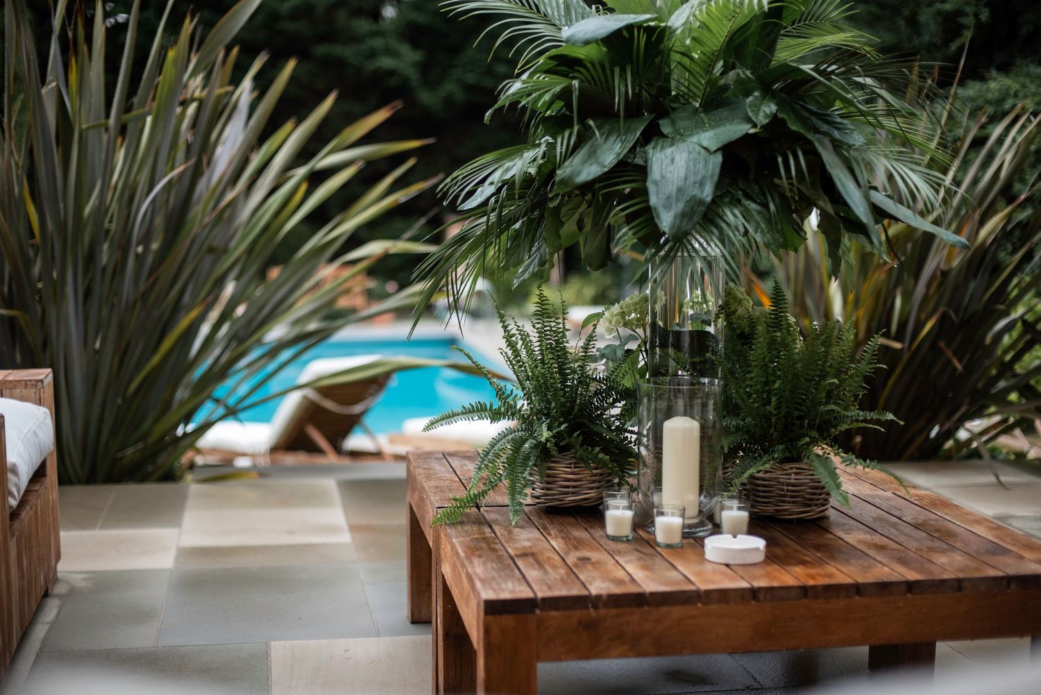 Wooden outdoor coffee table with tropical plants and white candles near a swimming pool.